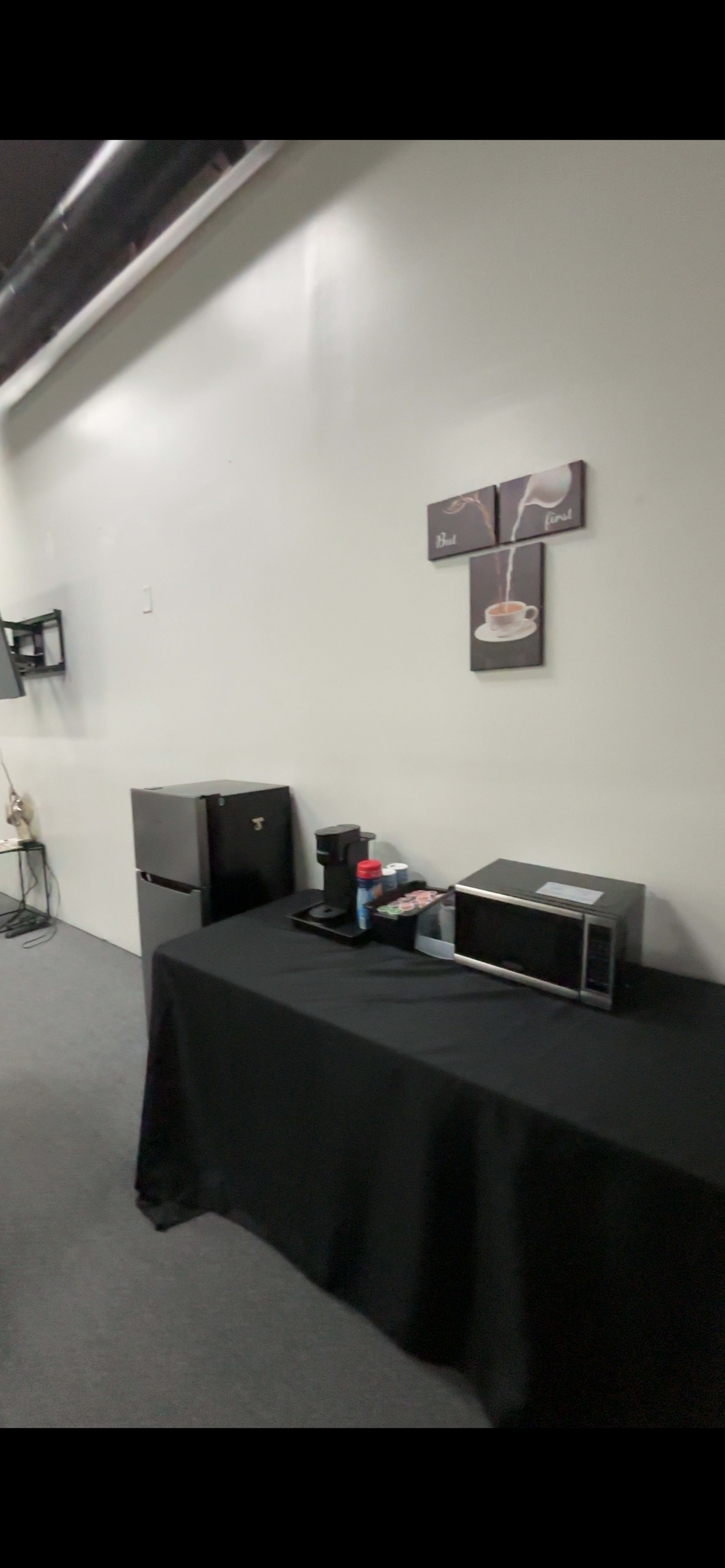The image shows a room corner with a black tablecloth covering a table that has a coffee maker, a microwave, and a small refrigerator against a plain white wall.