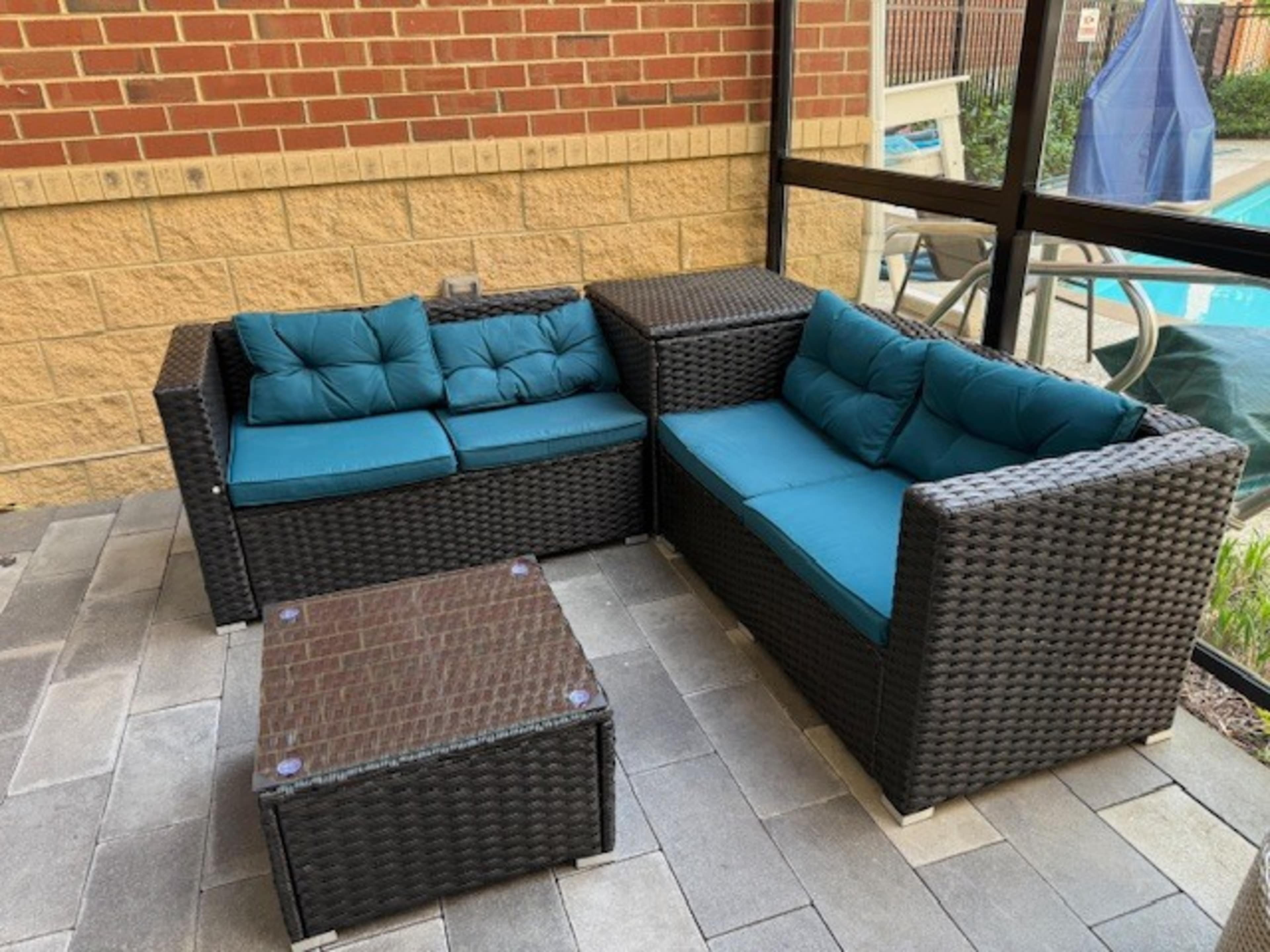 A wicker patio set with turquoise cushions is arranged beside a glass coffee table on a stone floor, with a pool visible in the background.