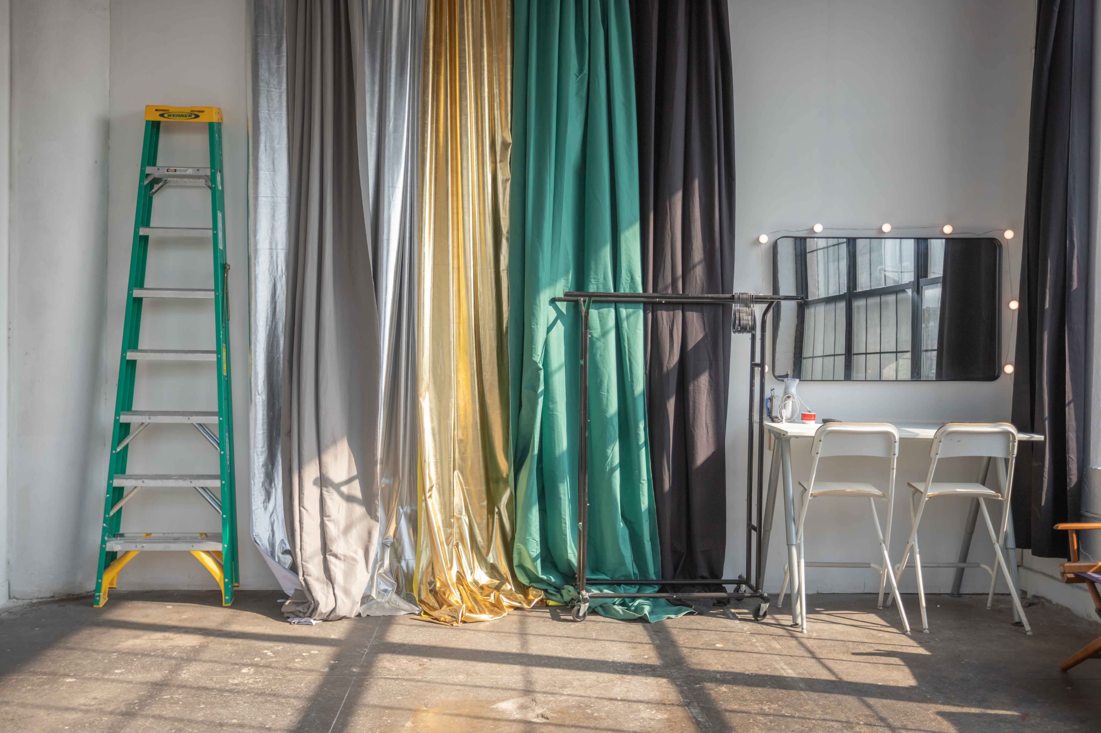 The image shows a studio space with a ladder, draped fabrics in various colors, a black clothing rack, and a mirror with light bulbs beside a set of white chairs.
