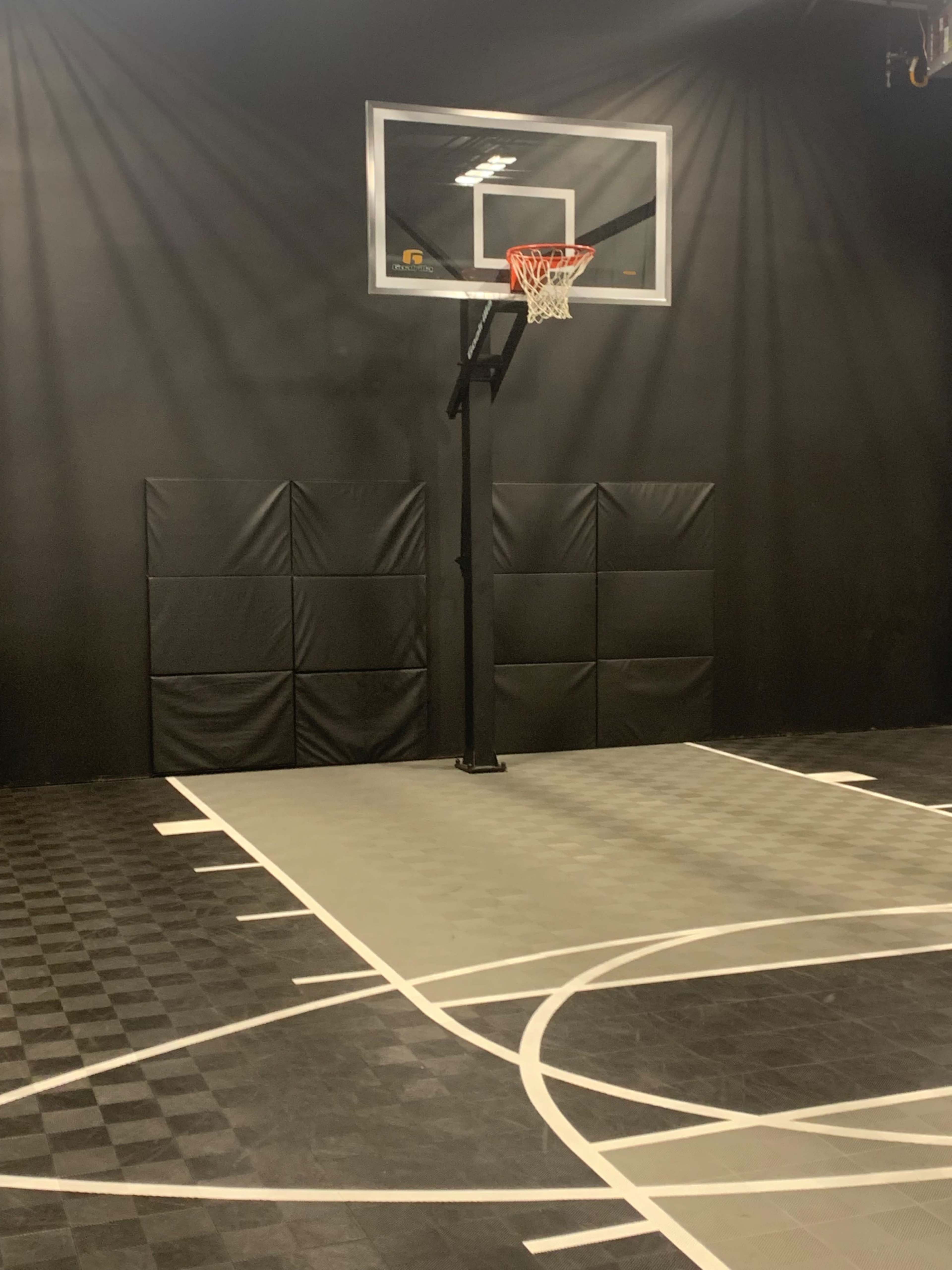 A basketball hoop mounted on a black wall, situated on a gym-style court with a gray floor and marked lines.
