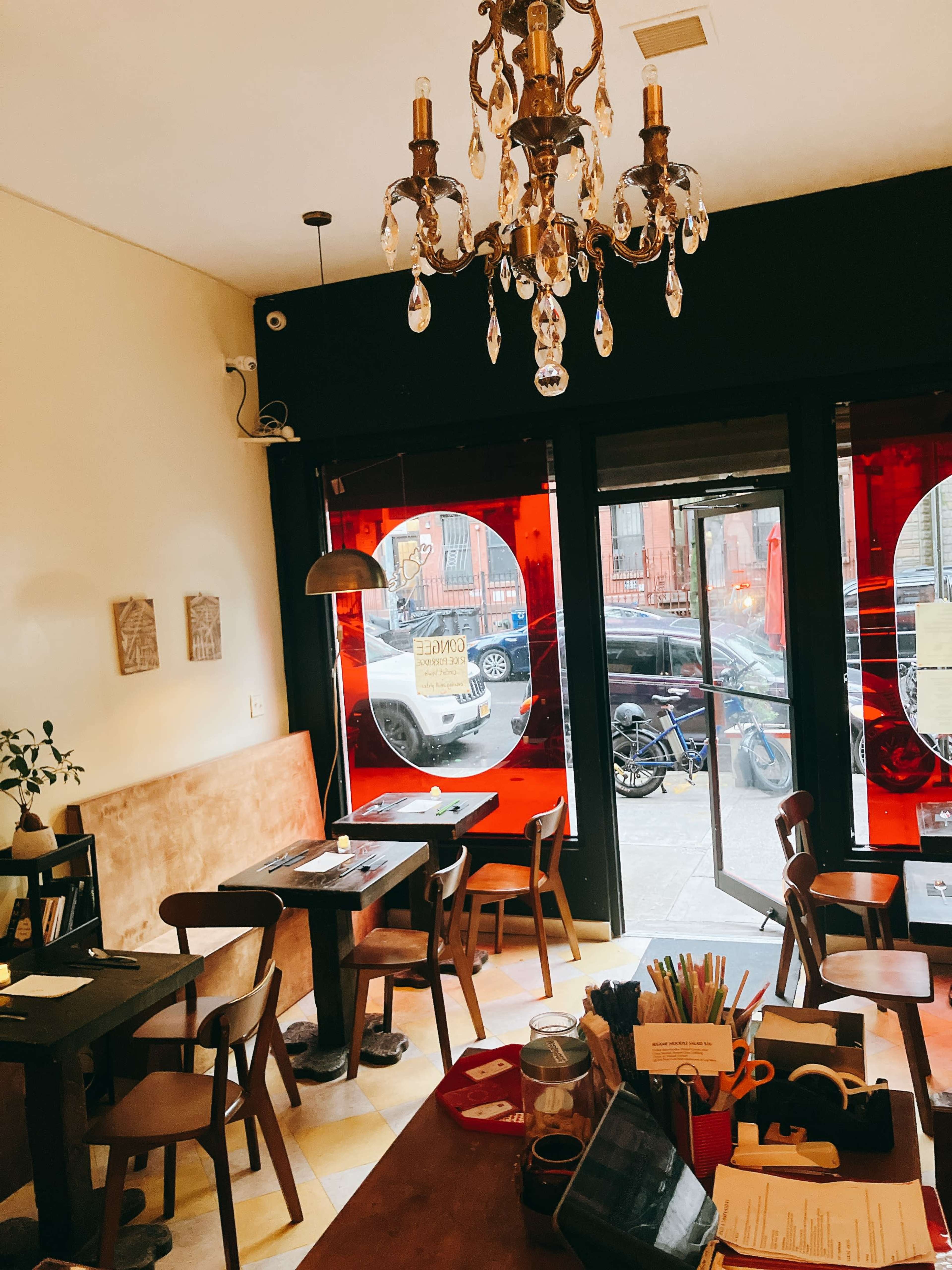 The image depicts a cozy cafe interior with wooden tables and chairs, a chandelier, and large windows featuring red glass panels.