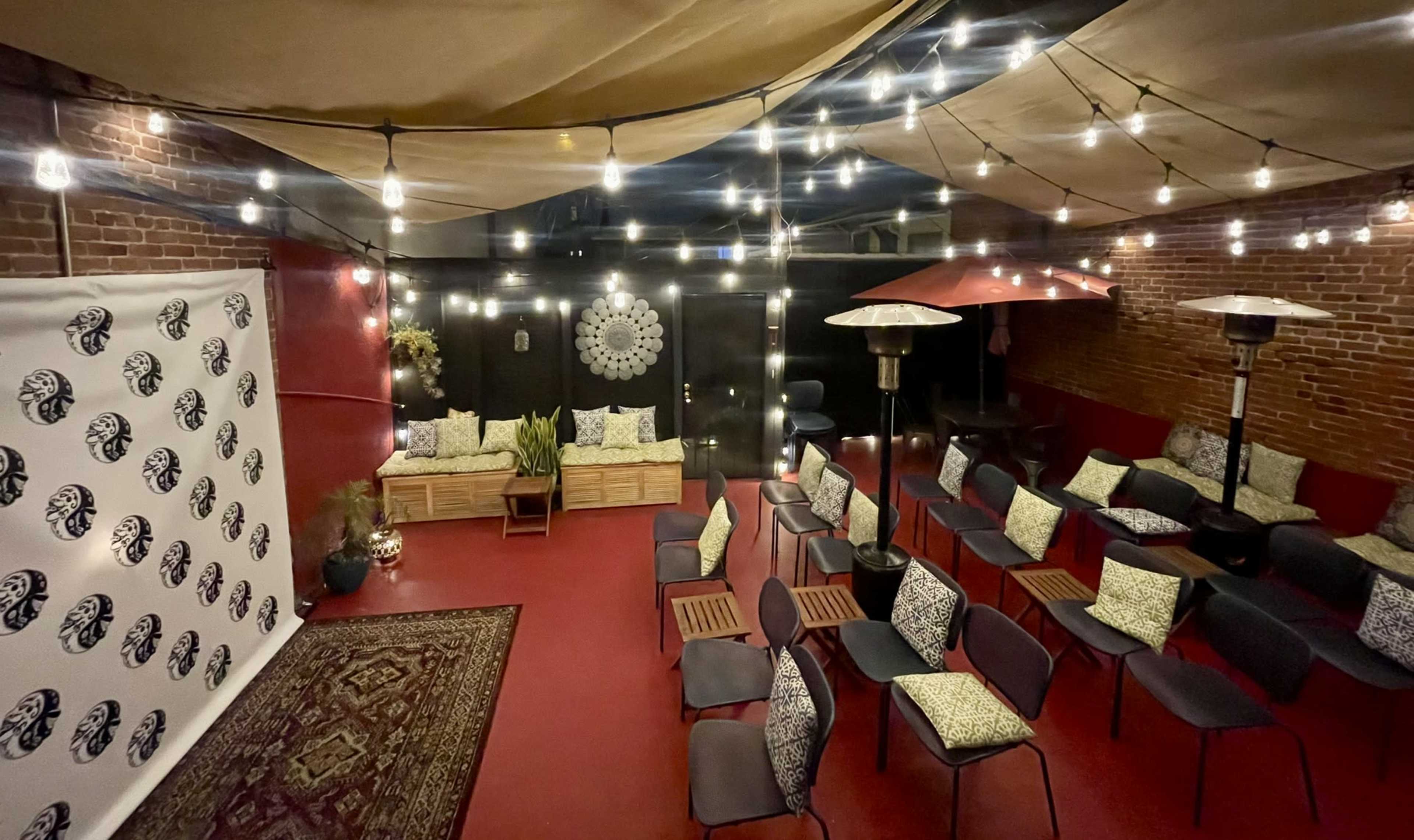 The image shows a cozy outdoor seating area with string lights overhead, arranged with chairs and small tables in front of a decorative wall.