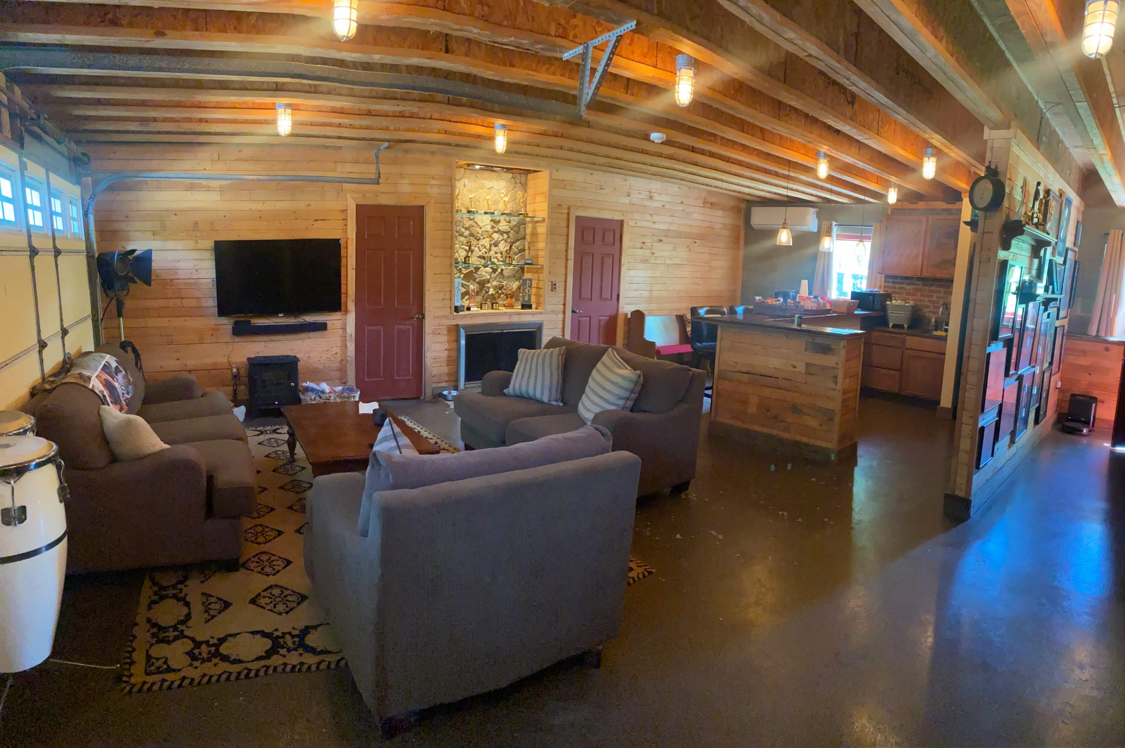 The image shows a cozy living space featuring a seating area with two sofas, a coffee table, a wall-mounted TV, and an open kitchen with wooden cabinetry and a stone wall accent.