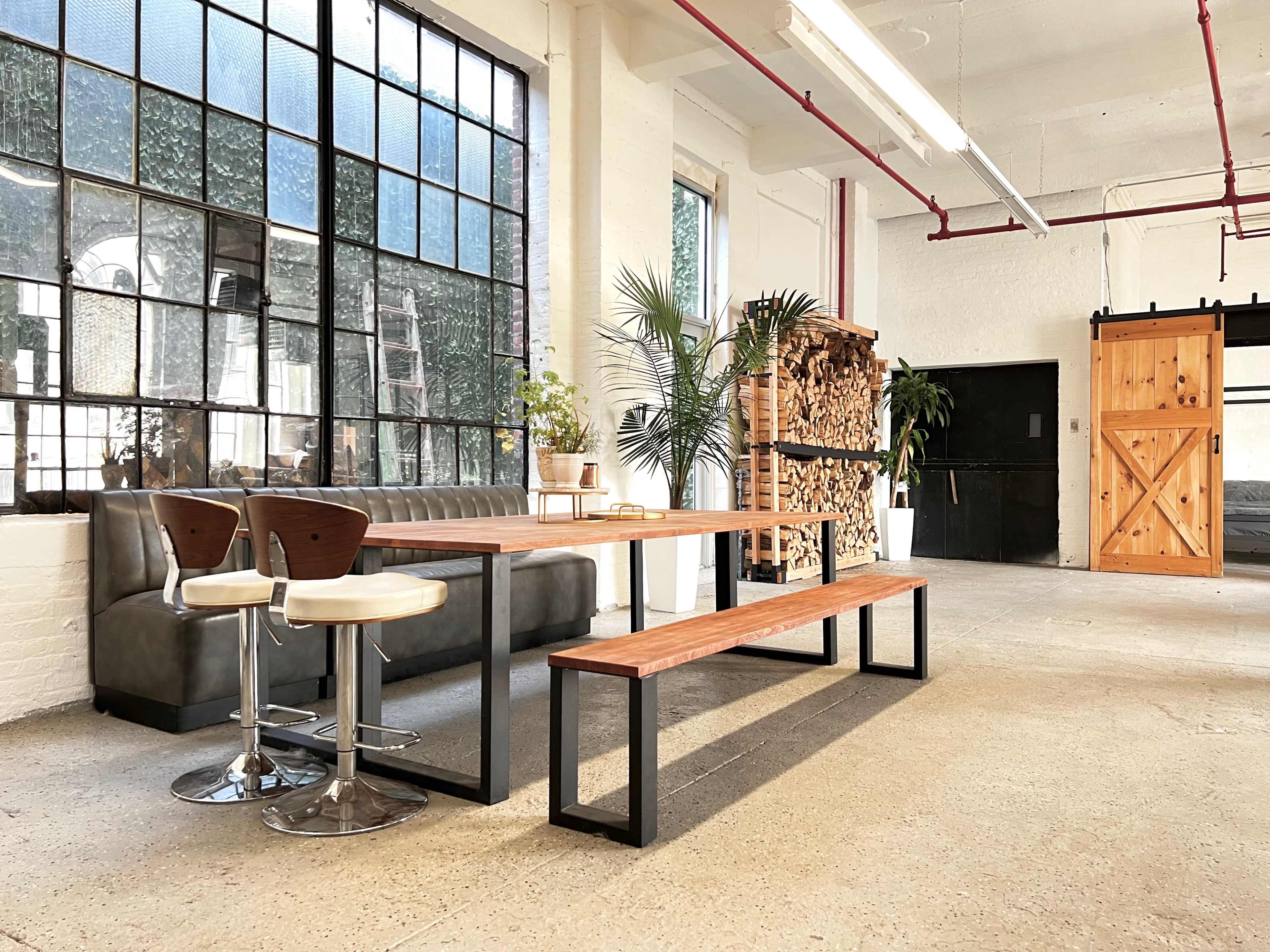 A modern interior space features a long wooden table with benches, a black leather couch, and large windows that allow natural light to enter.