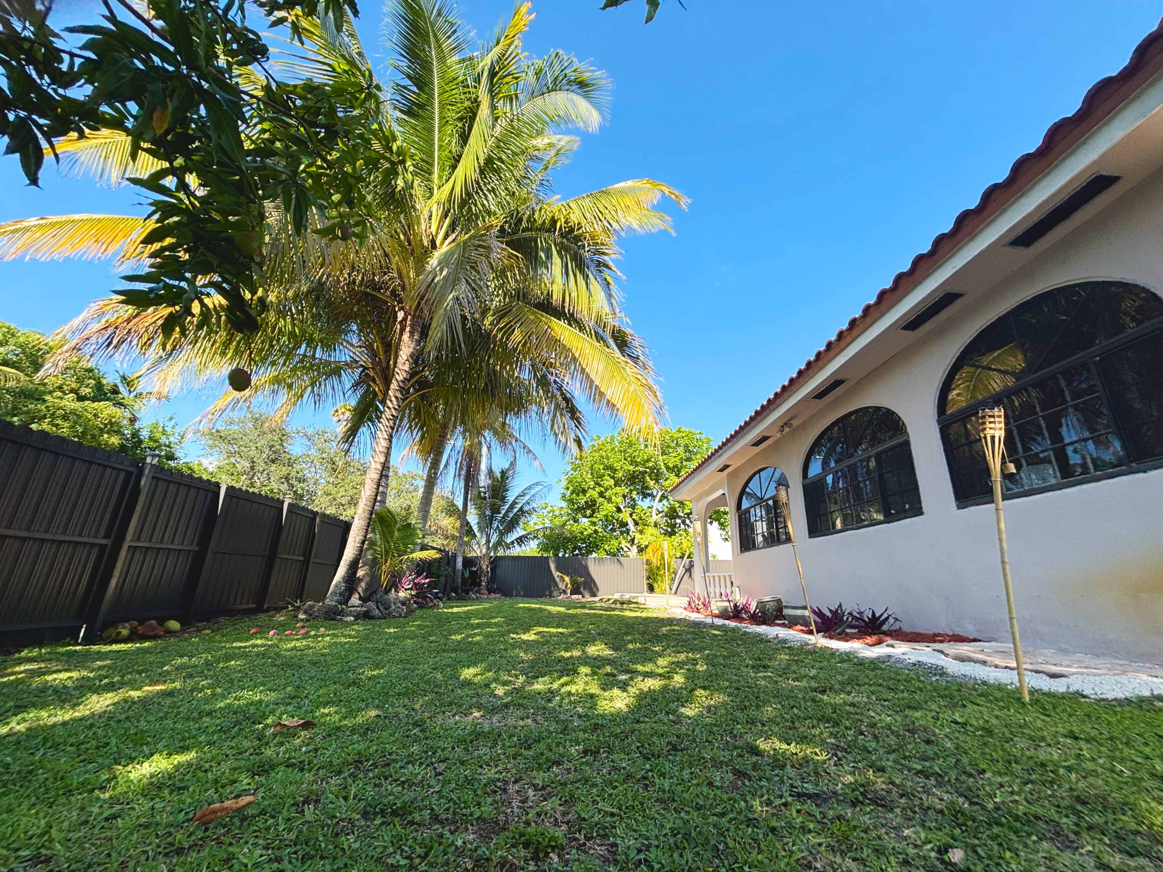 The image shows a well-maintained backyard with a lawn, palm tree, and a house featuring arched windows.