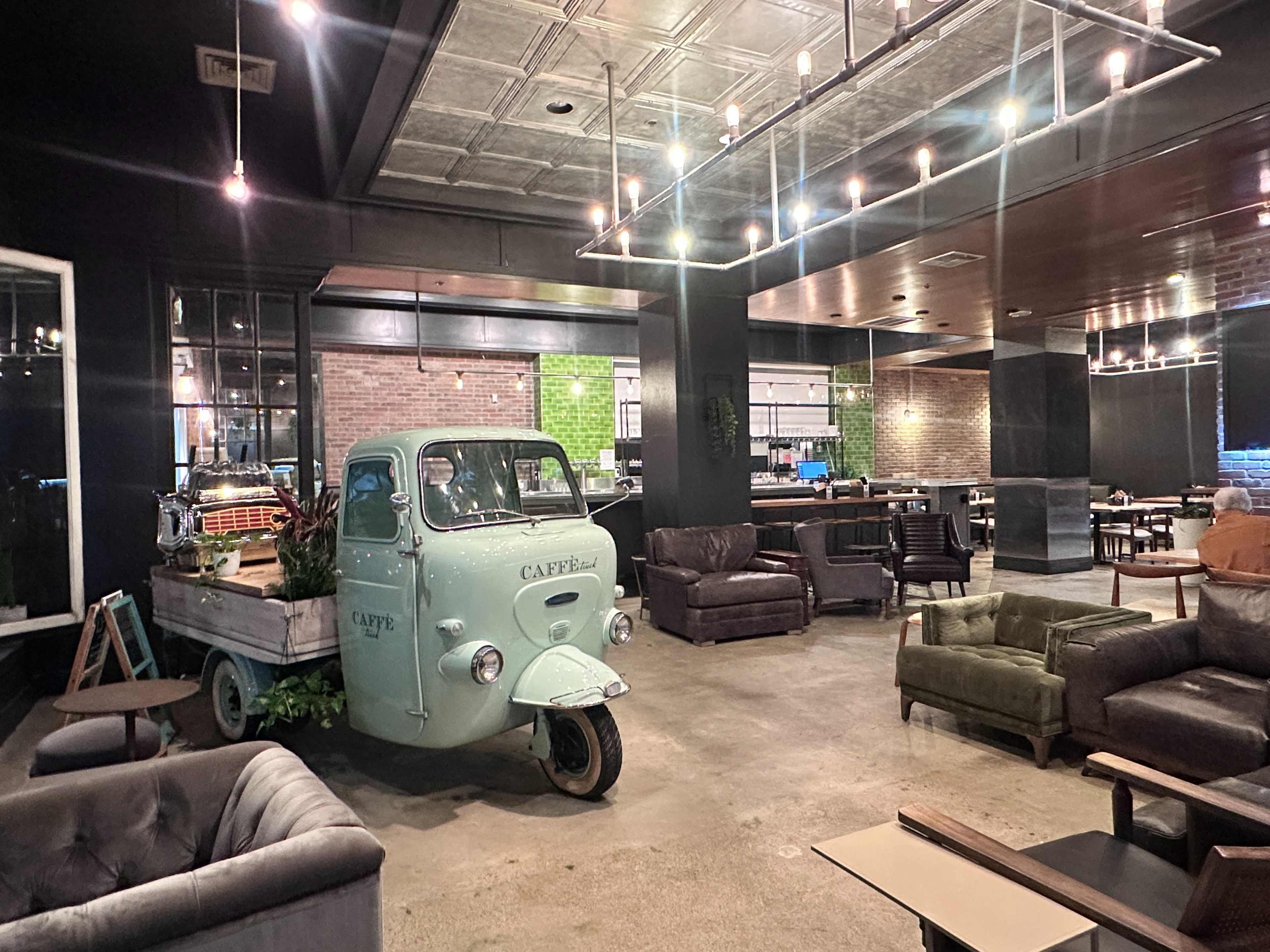 The image shows a modern café interior featuring a mint-green vintage delivery vehicle filled with plants, surrounded by various seating arrangements in a spacious setting.