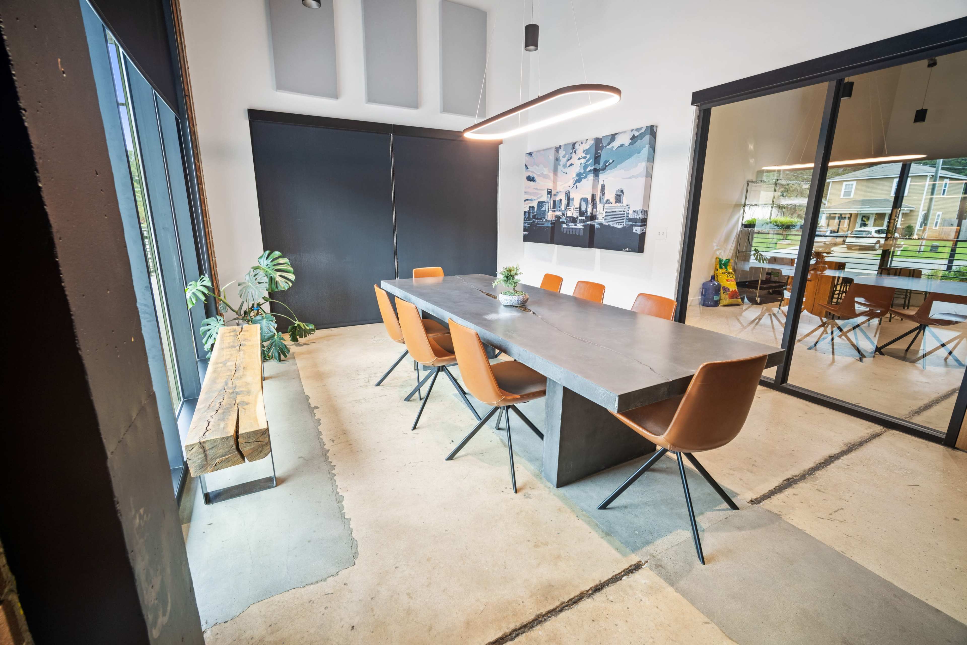 A modern meeting room features a long concrete table surrounded by leather chairs, with large windows and a wall-mounted artwork.