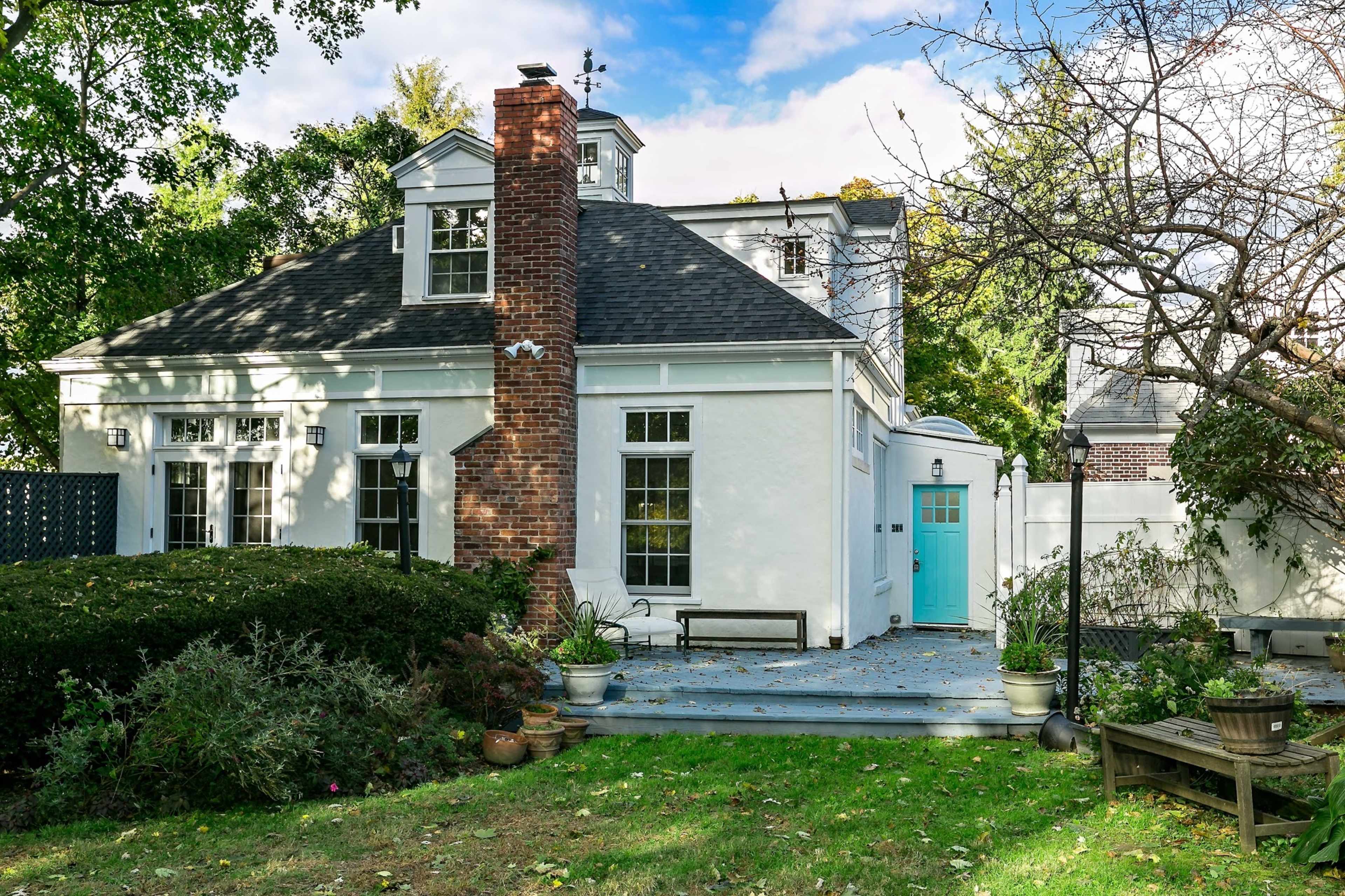A white house with a brick chimney features a blue door and is surrounded by a well-maintained yard with trees and shrubs.