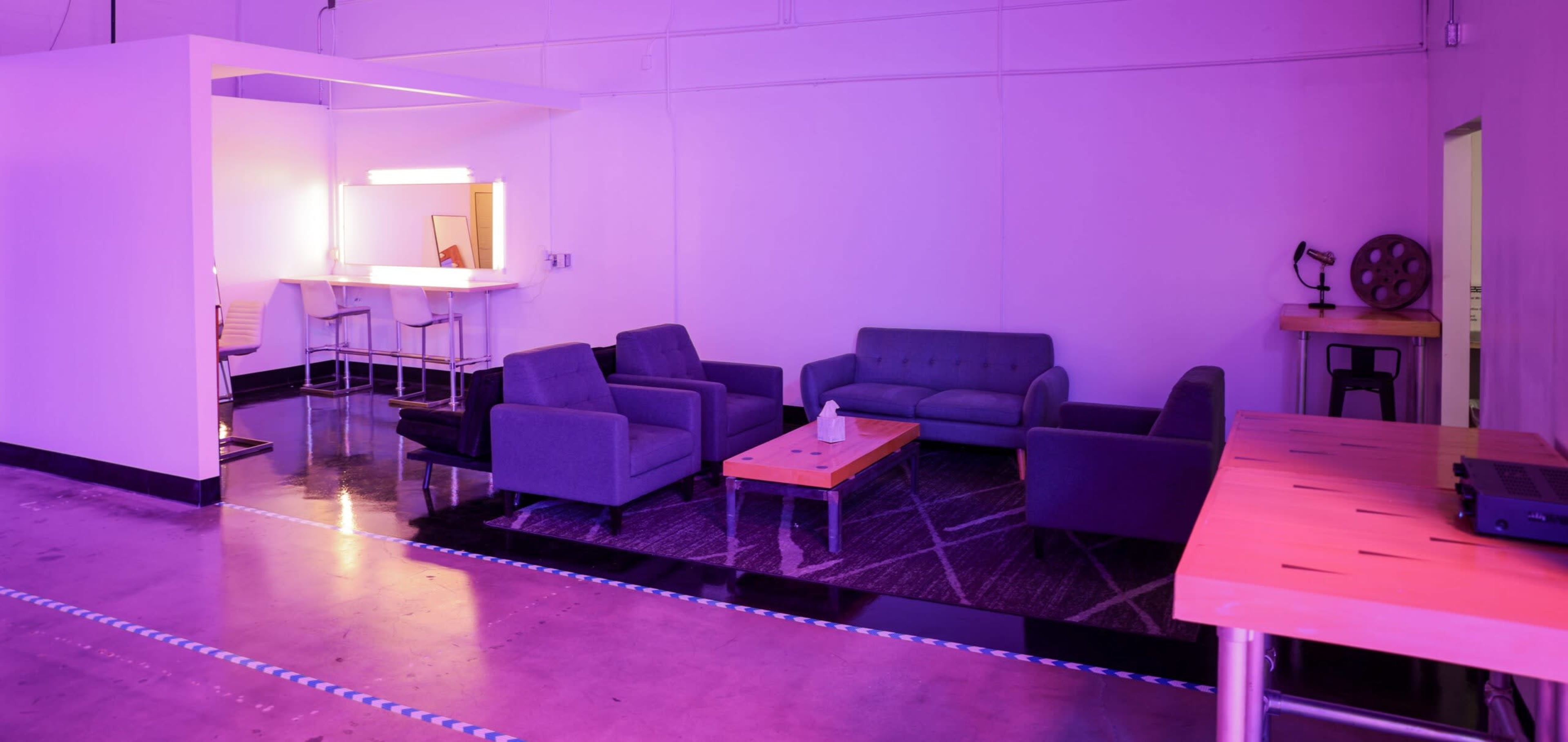 A modern, empty lounge area featuring gray sofas, a wooden coffee table, a bar counter with stools, and ambient purple lighting.