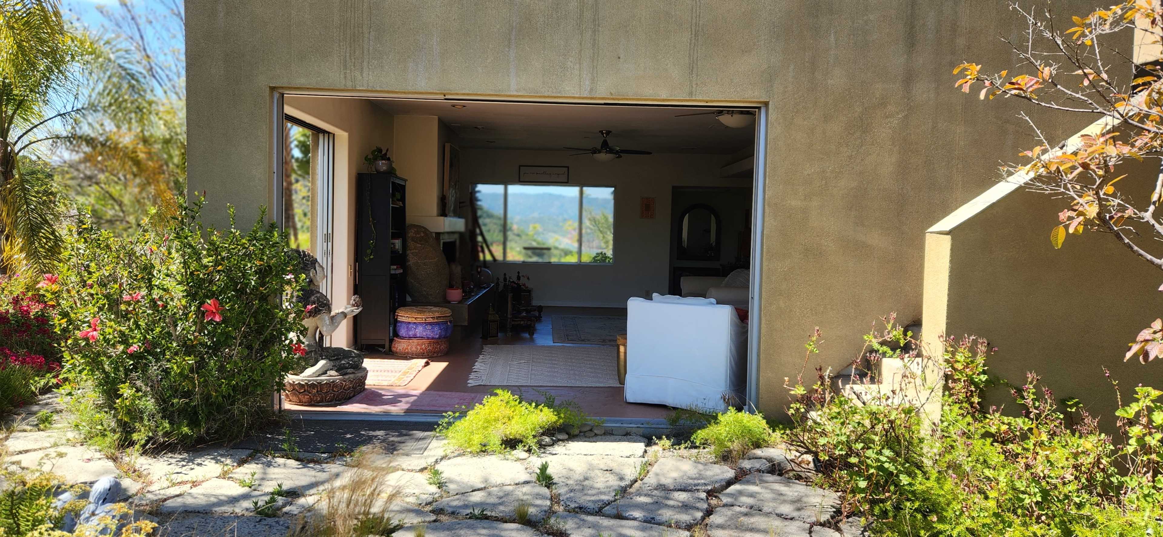 An open doorway of a house with a view of a green landscape, framed by plants and stone pathways.