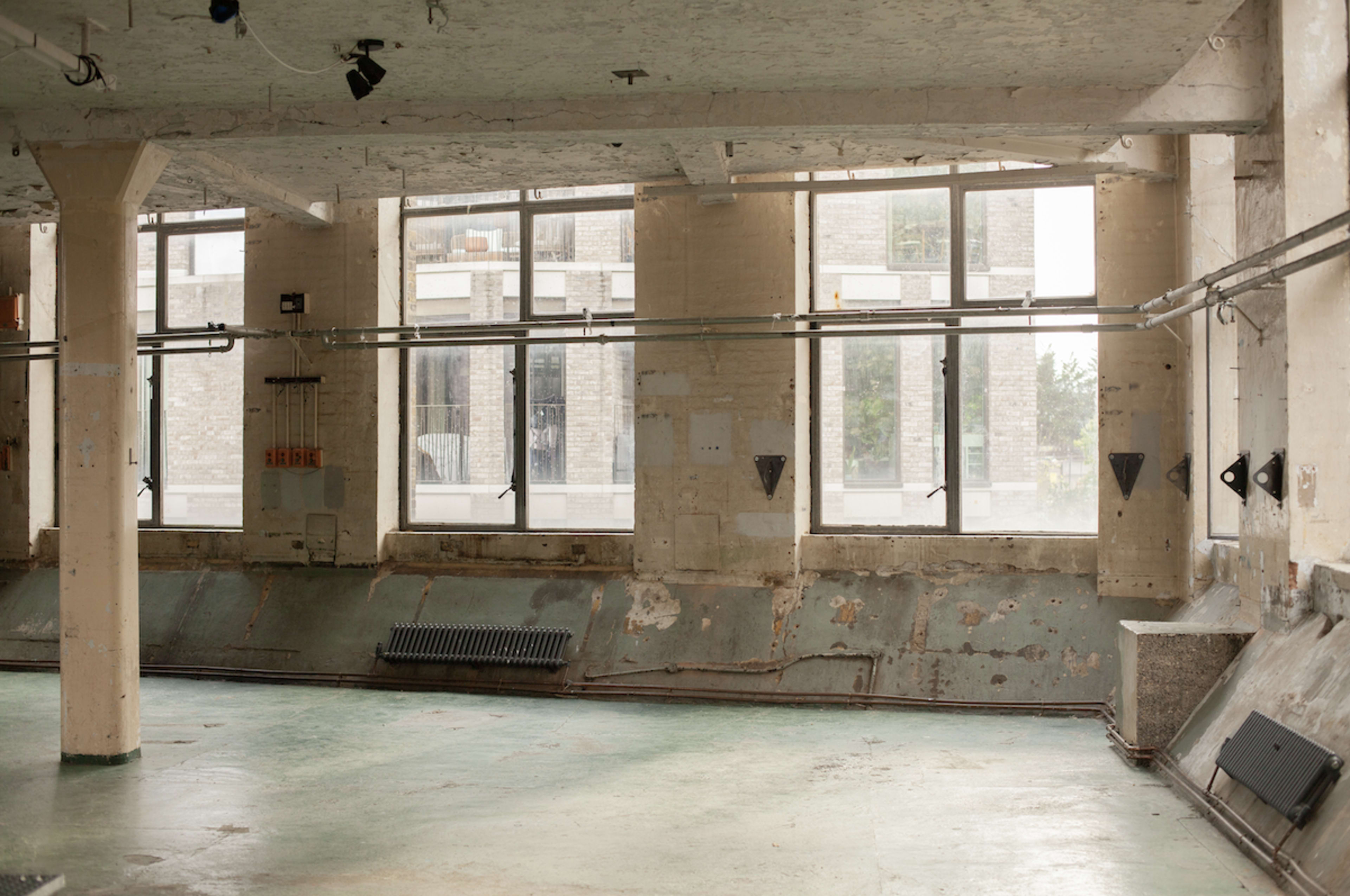 The image shows an empty industrial space with large windows, peeling walls, and a green floor.