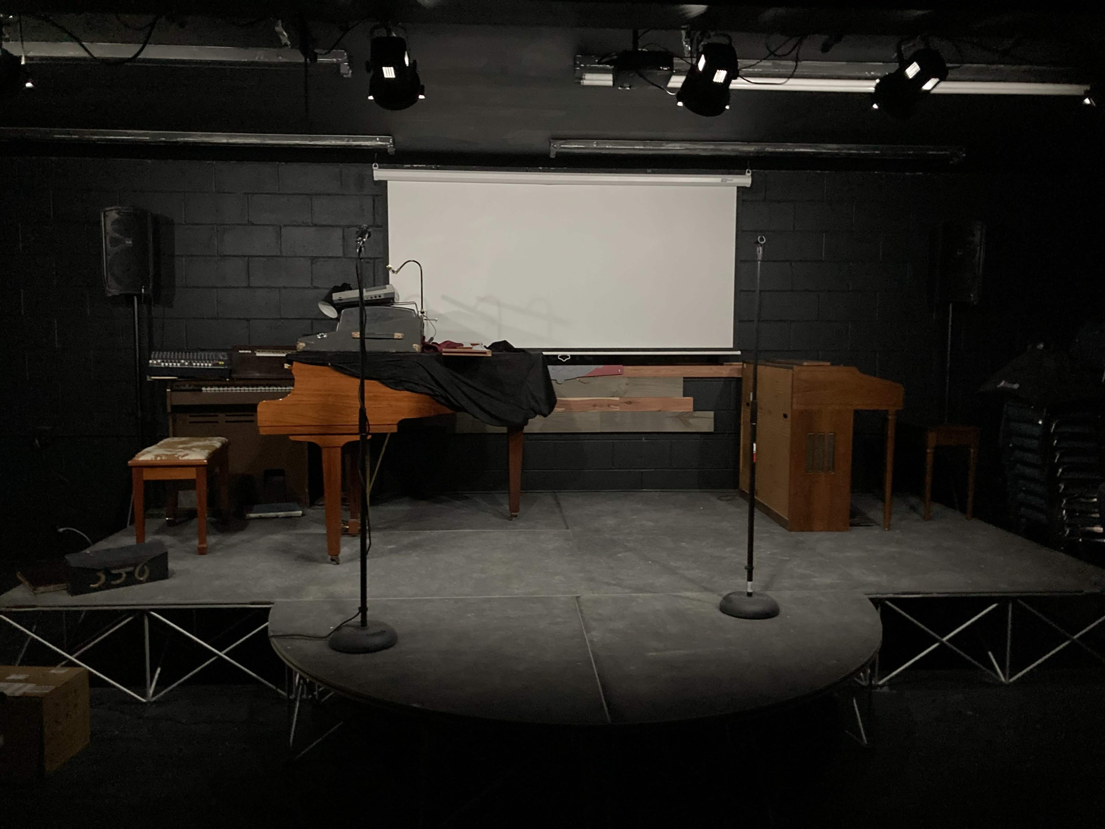 A stage setup with a grand piano, an organ, and microphones, against a black backdrop with a projection screen.