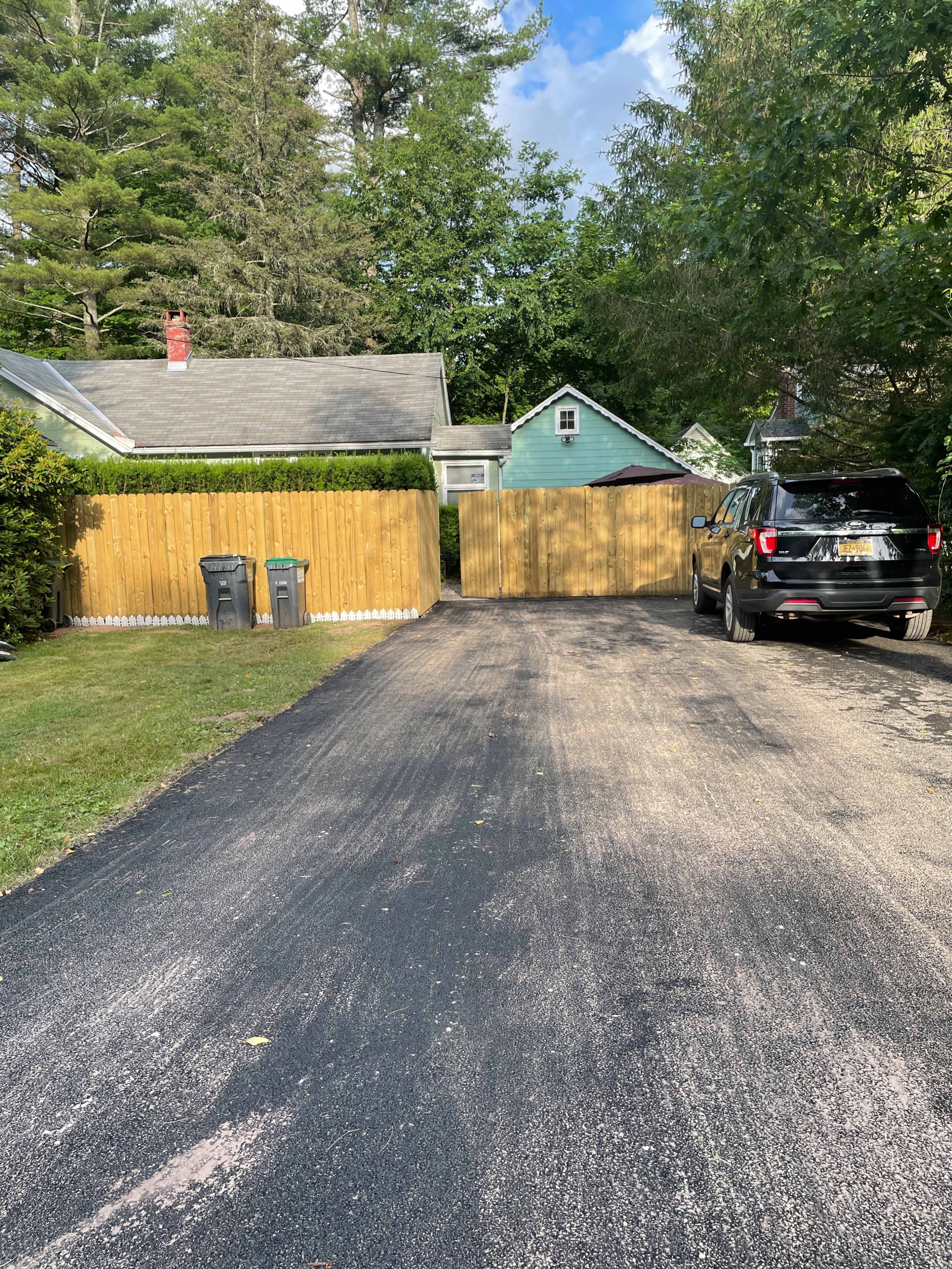 The image shows a driveway leading up to a wooden gate, flanked by two trash bins and a black SUV parked on the right side.