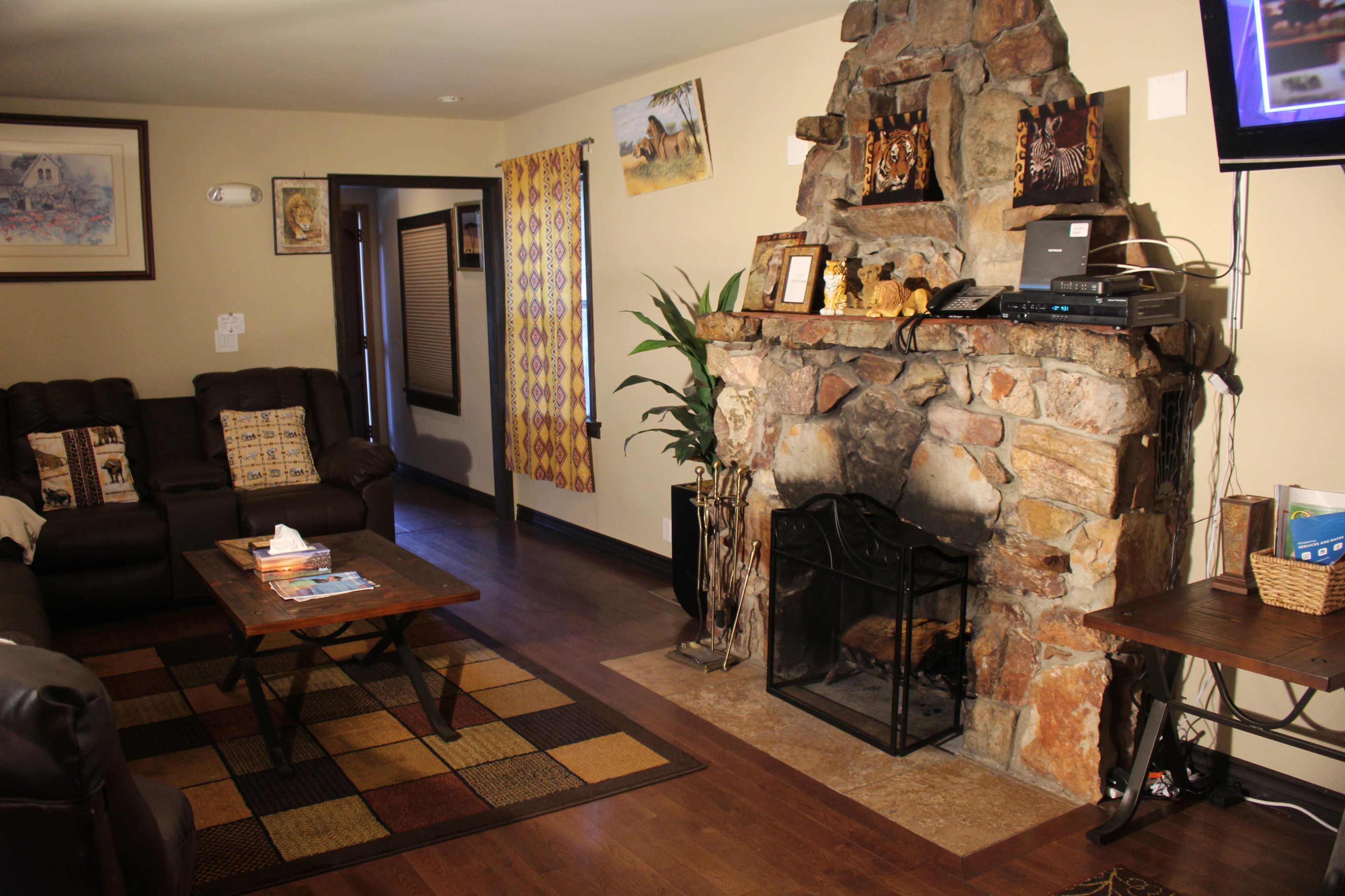 The image shows a living room featuring a stone fireplace, two leather sofas, a coffee table, and decorative elements on the walls.