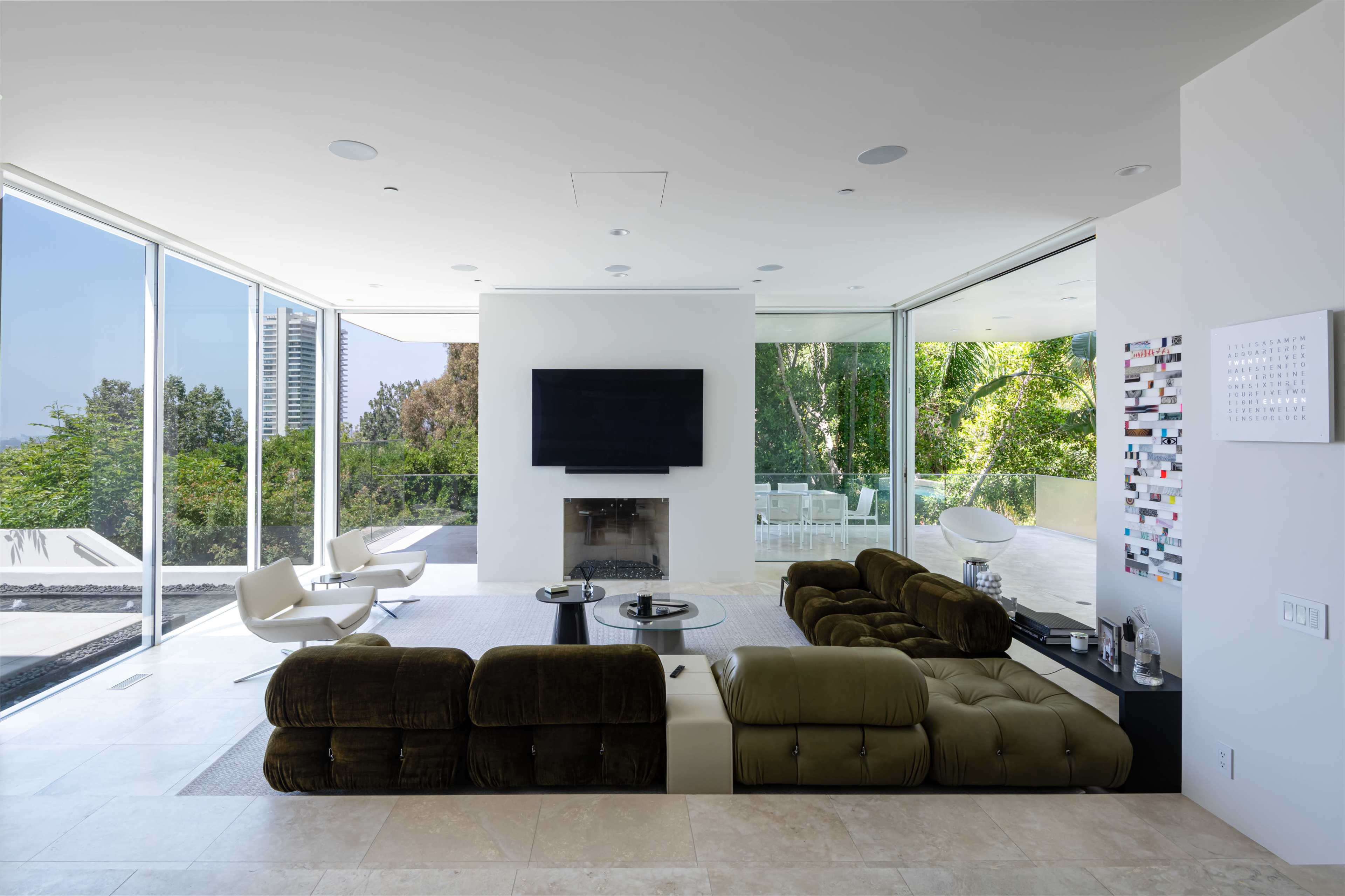 A modern living room features a sectional sofa, a coffee table, a fireplace, and large glass walls that provide views of the outdoors.