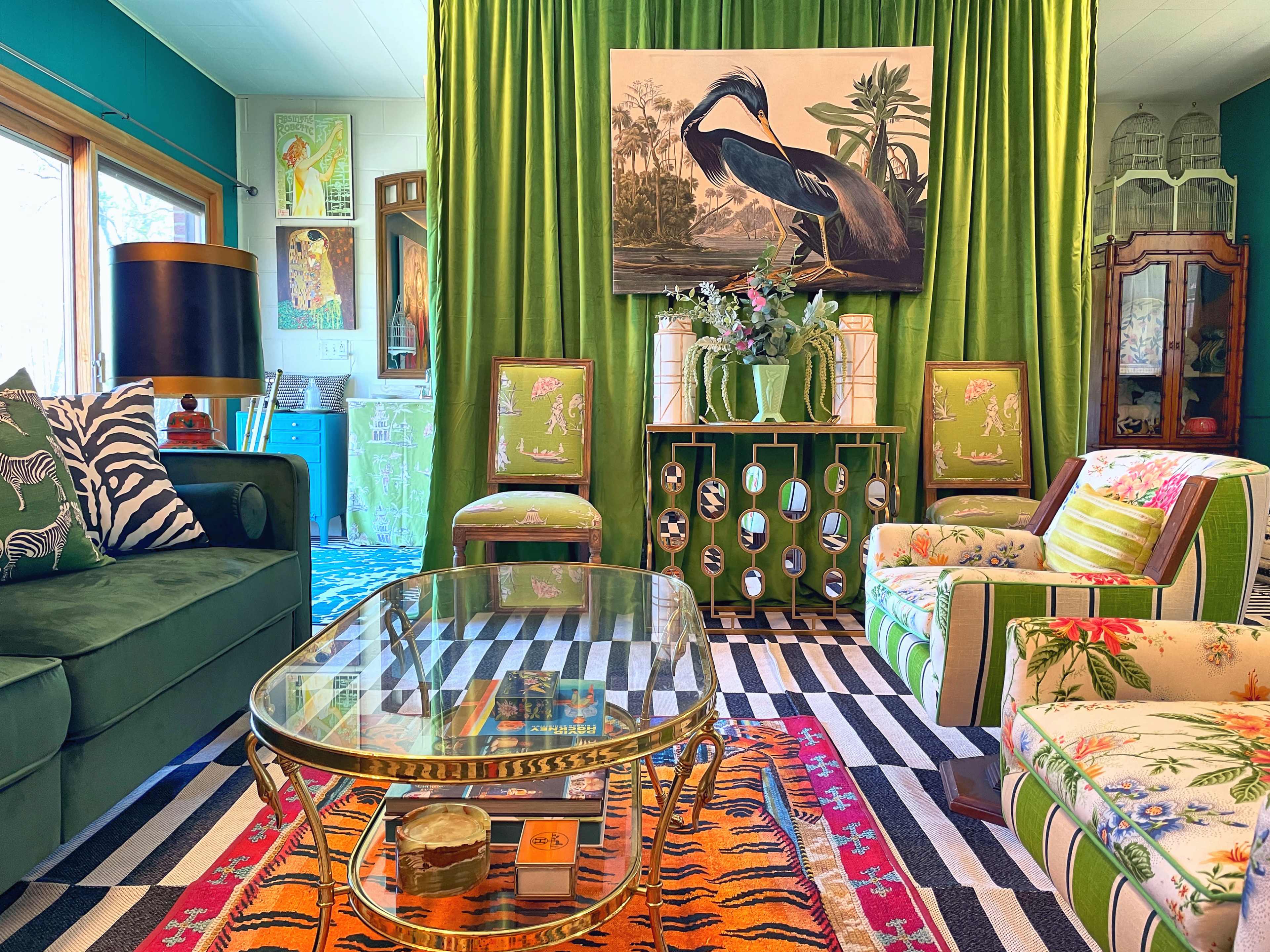 A colorful living room featuring a green velvet sofa, a glass coffee table, striped rugs, and vibrant artwork on the walls, with a large painting of a bird above a console table adorned with flowers.