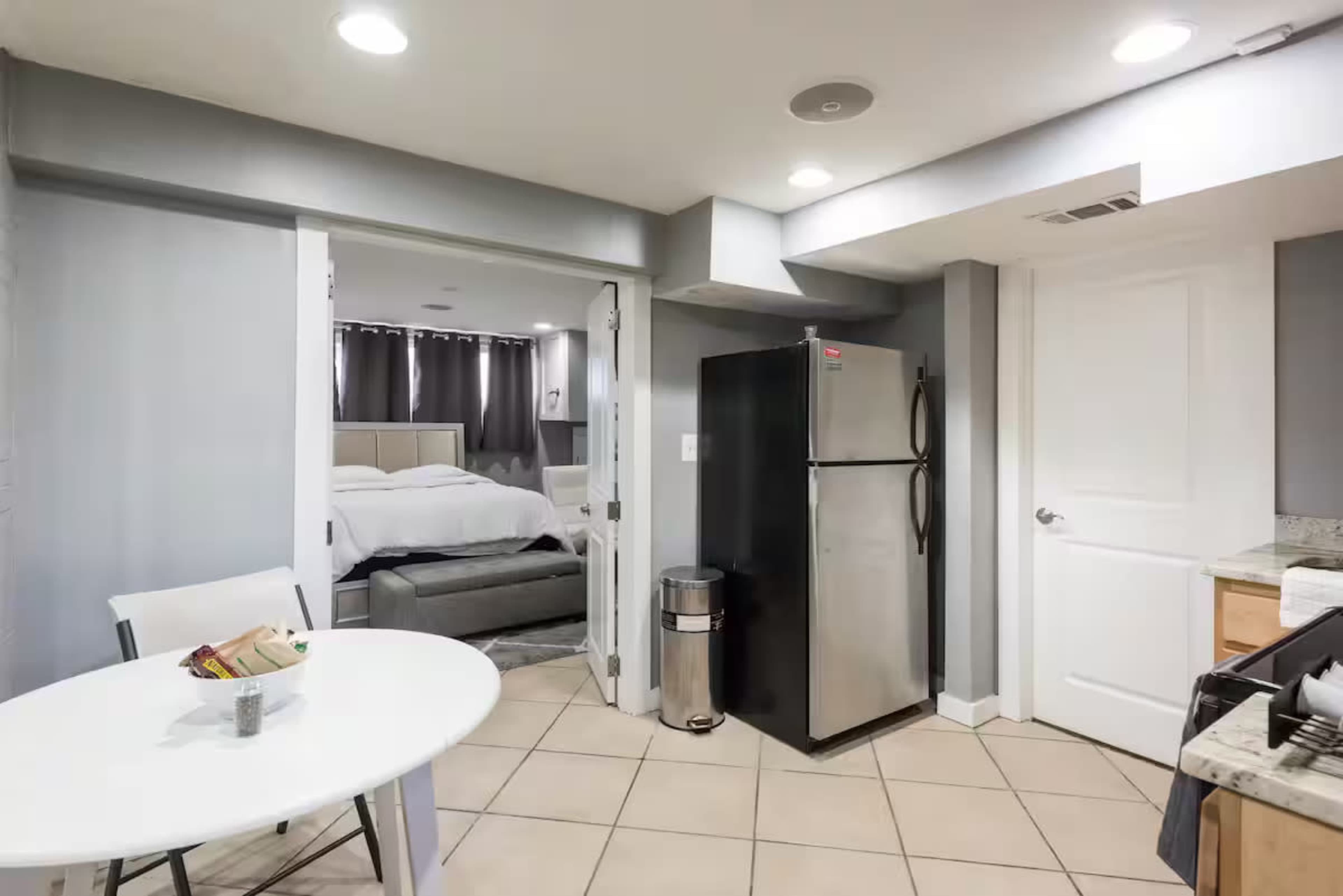 The image shows a small apartment featuring a kitchen area with a refrigerator and a round dining table, adjacent to a living space with a bed visible through an open door.