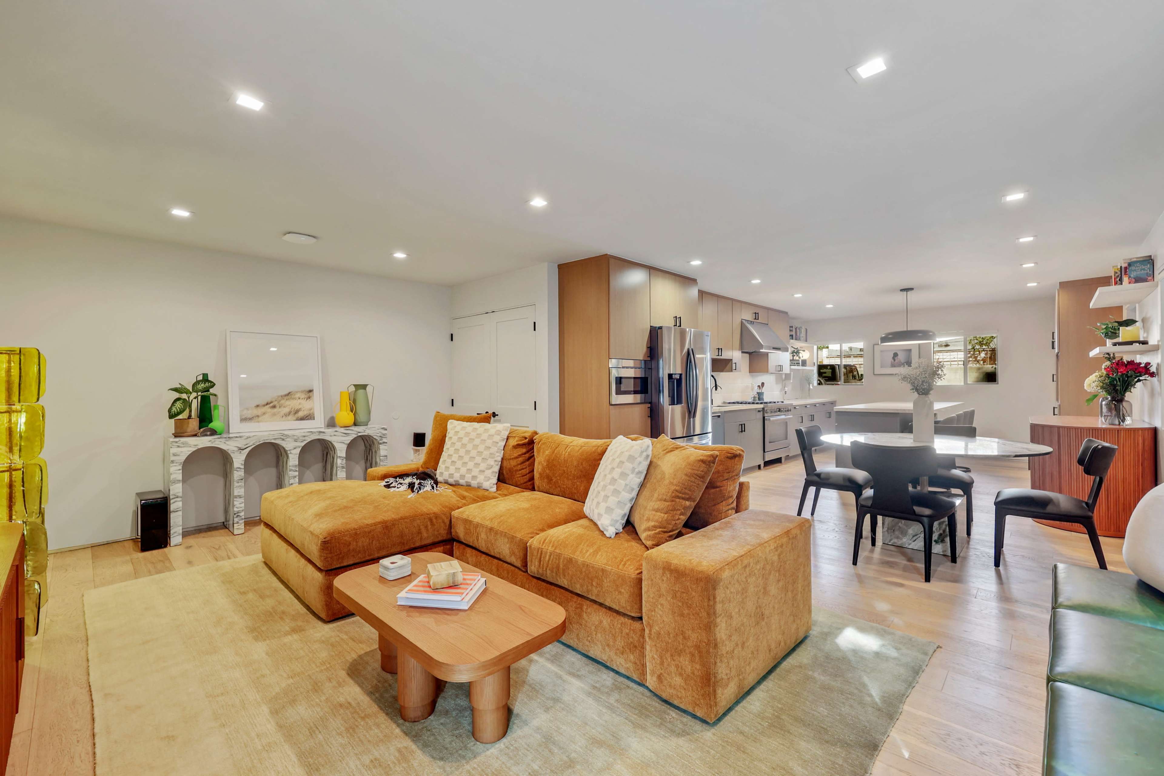 The image shows a modern living space with a large brown sectional sofa, a wooden coffee table, and a dining area featuring a round table and black chairs, all illuminated by ceiling lights.