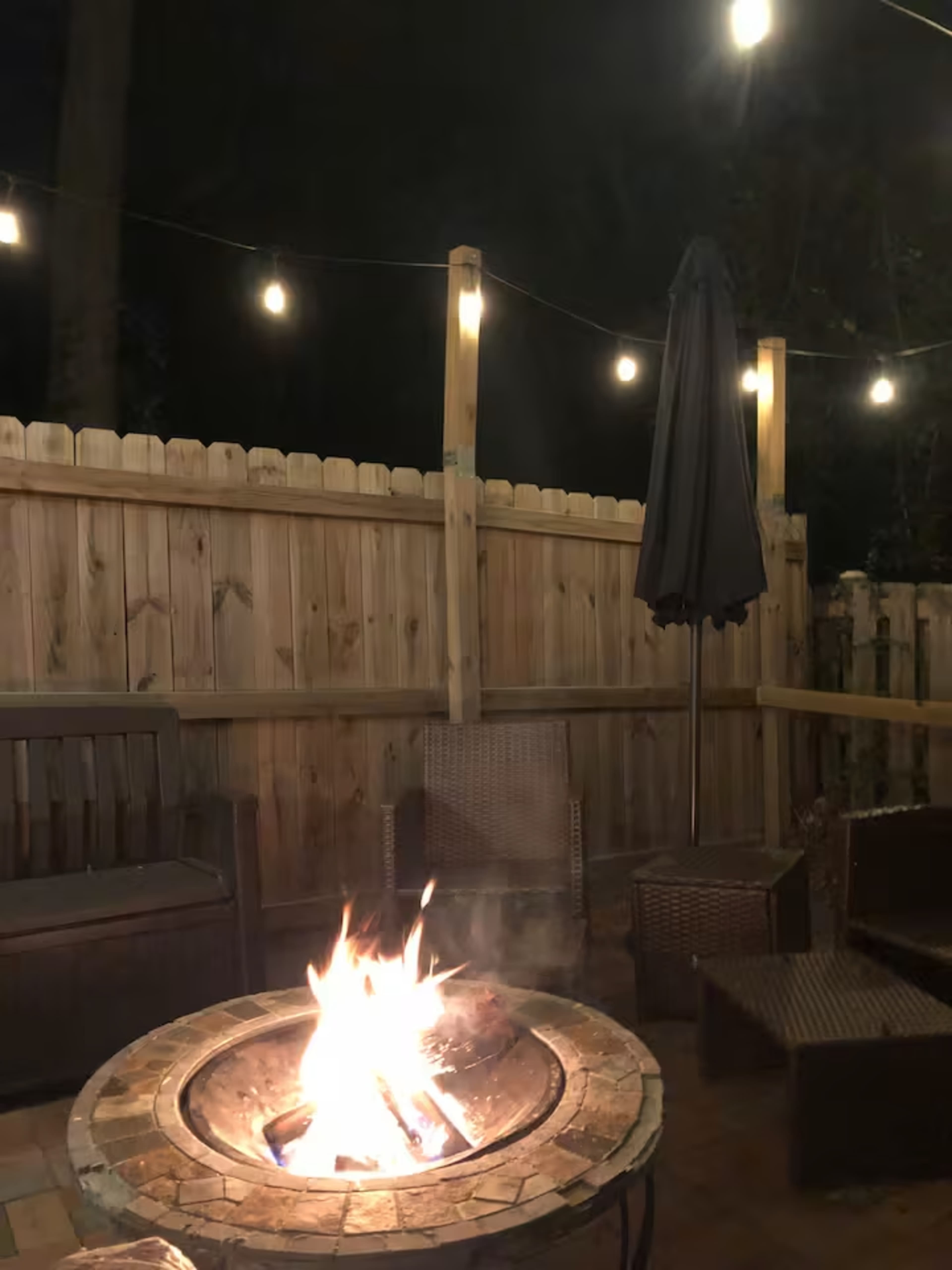 A fire pit burns brightly in a patio area, surrounded by woven chairs and illuminated by string lights above.