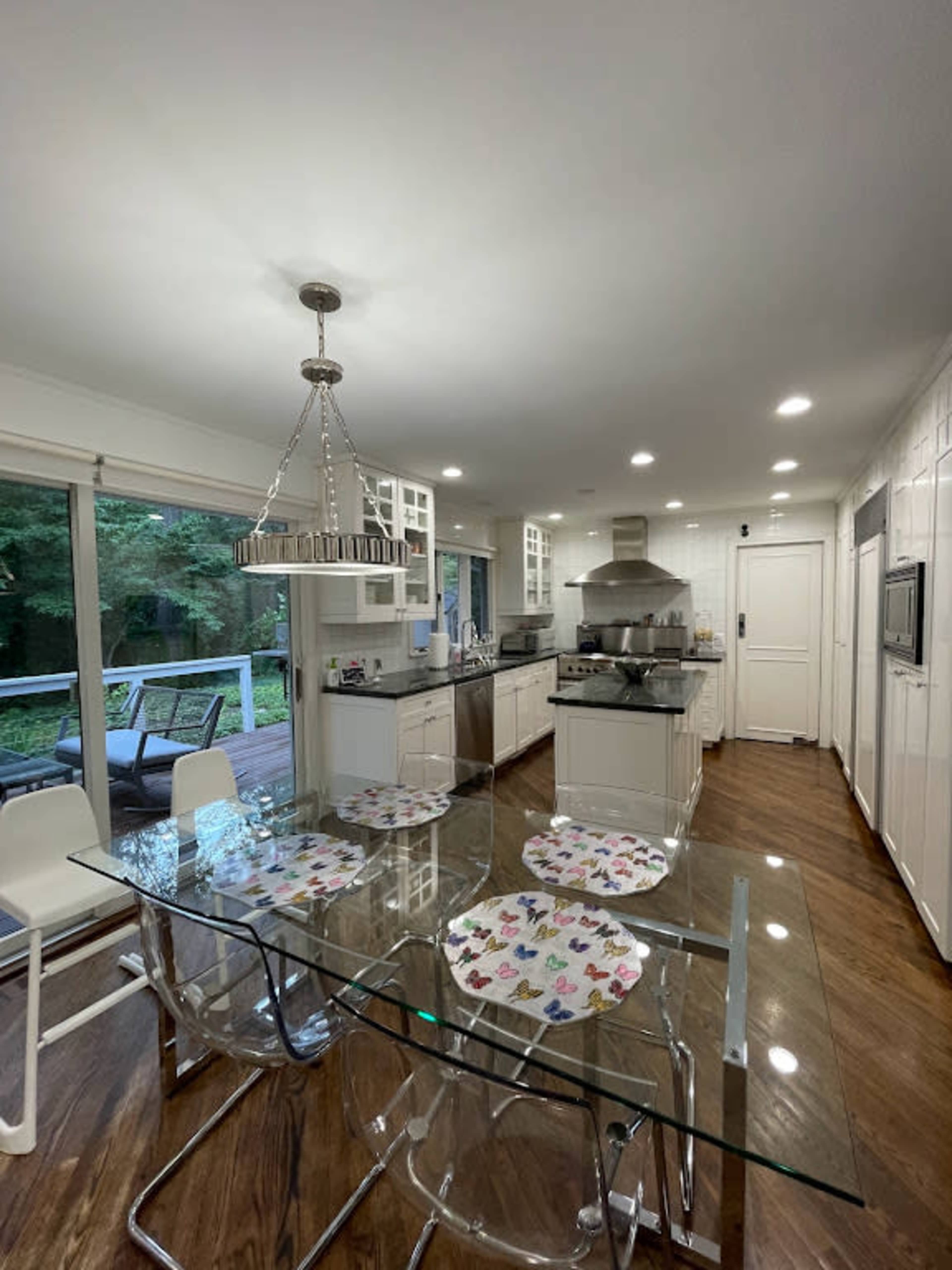 The image depicts a modern kitchen with a glass dining table and a view of a deck surrounded by trees.