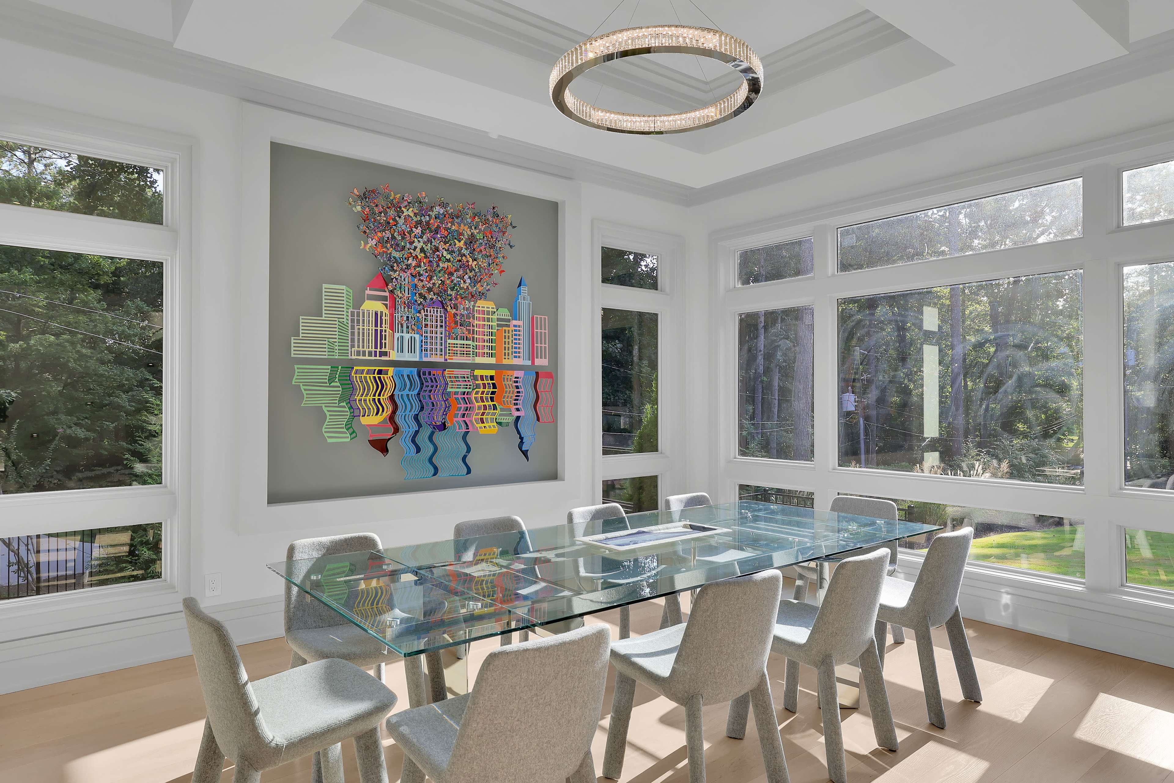 A modern dining room features a glass table surrounded by eight light-colored chairs, with large windows providing a view of greenery and a colorful wall art piece depicting stylized city buildings.