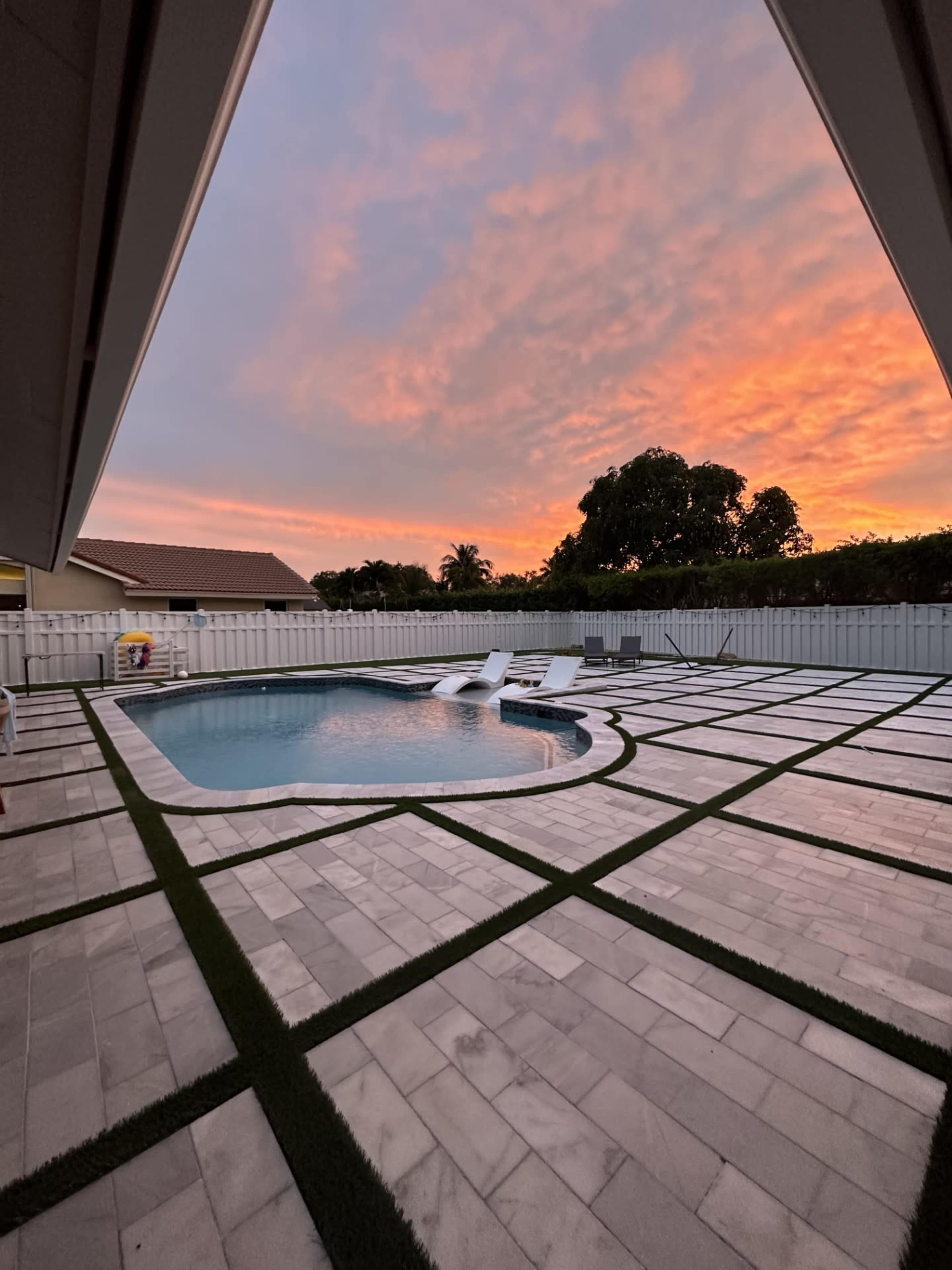 The image shows a swimming pool surrounded by a paved patio and grass patches, with a colorful sunset in the background.