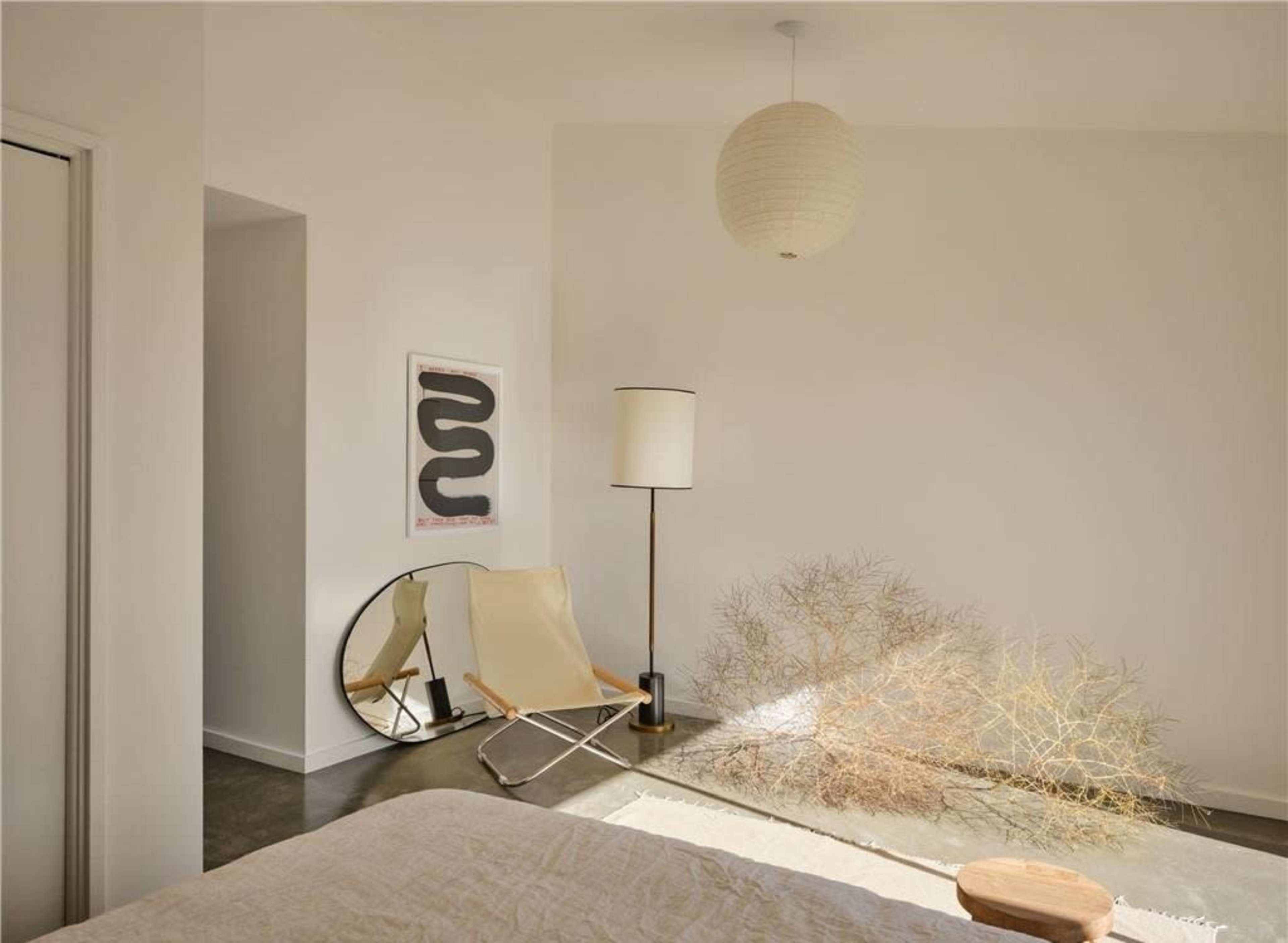 A minimalist bedroom with a bed, a floor lamp, a chair, a decorative mirror, and a dried plant arrangement in a sunlit corner.