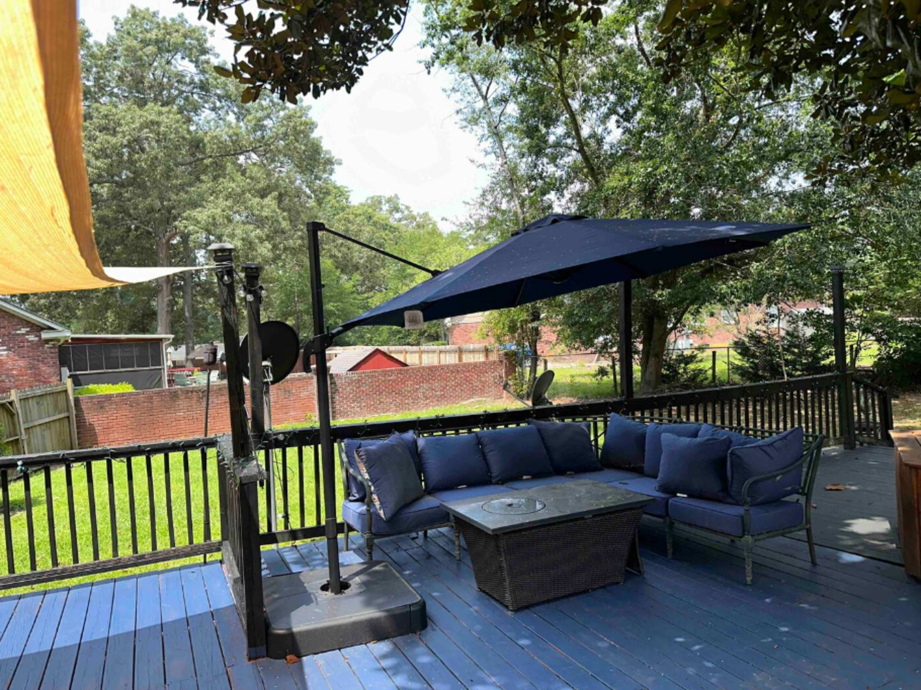 A shaded outdoor seating area features a blue sofa set and a table on a wooden deck with a green backyard in the background.