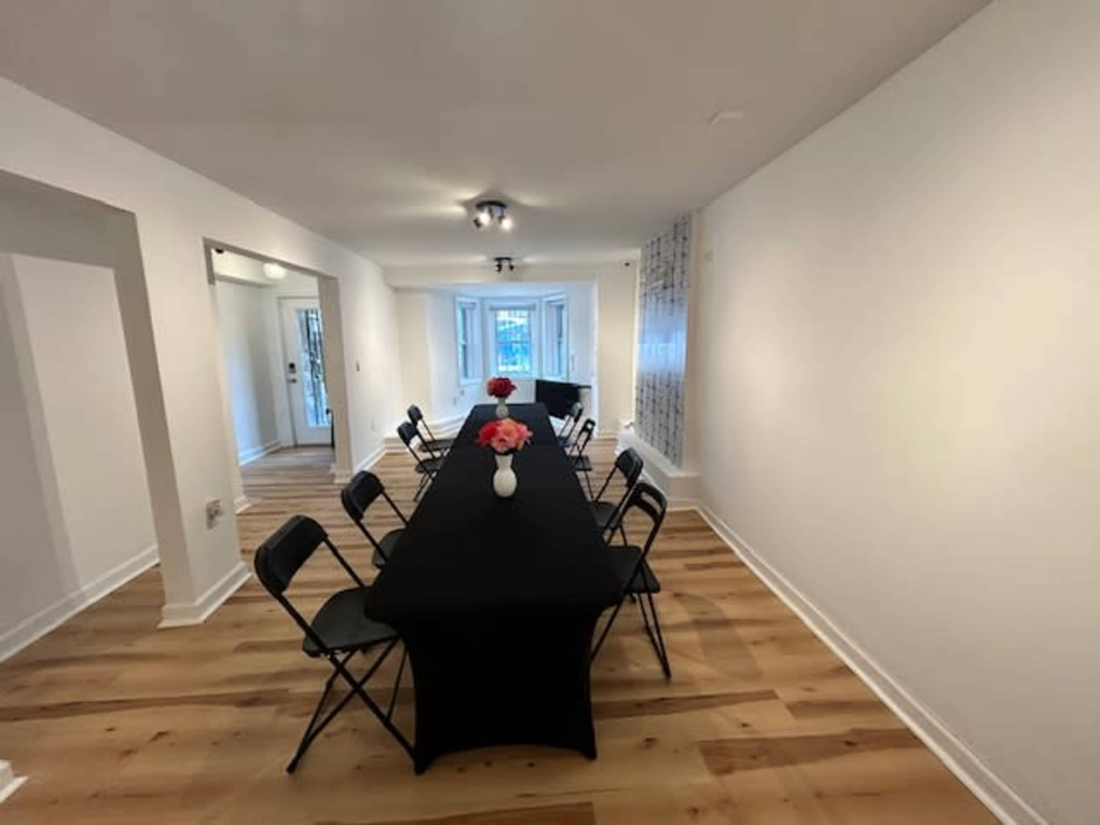 A long black table with chairs is set up in a bright, spacious room adorned with a flower vase.