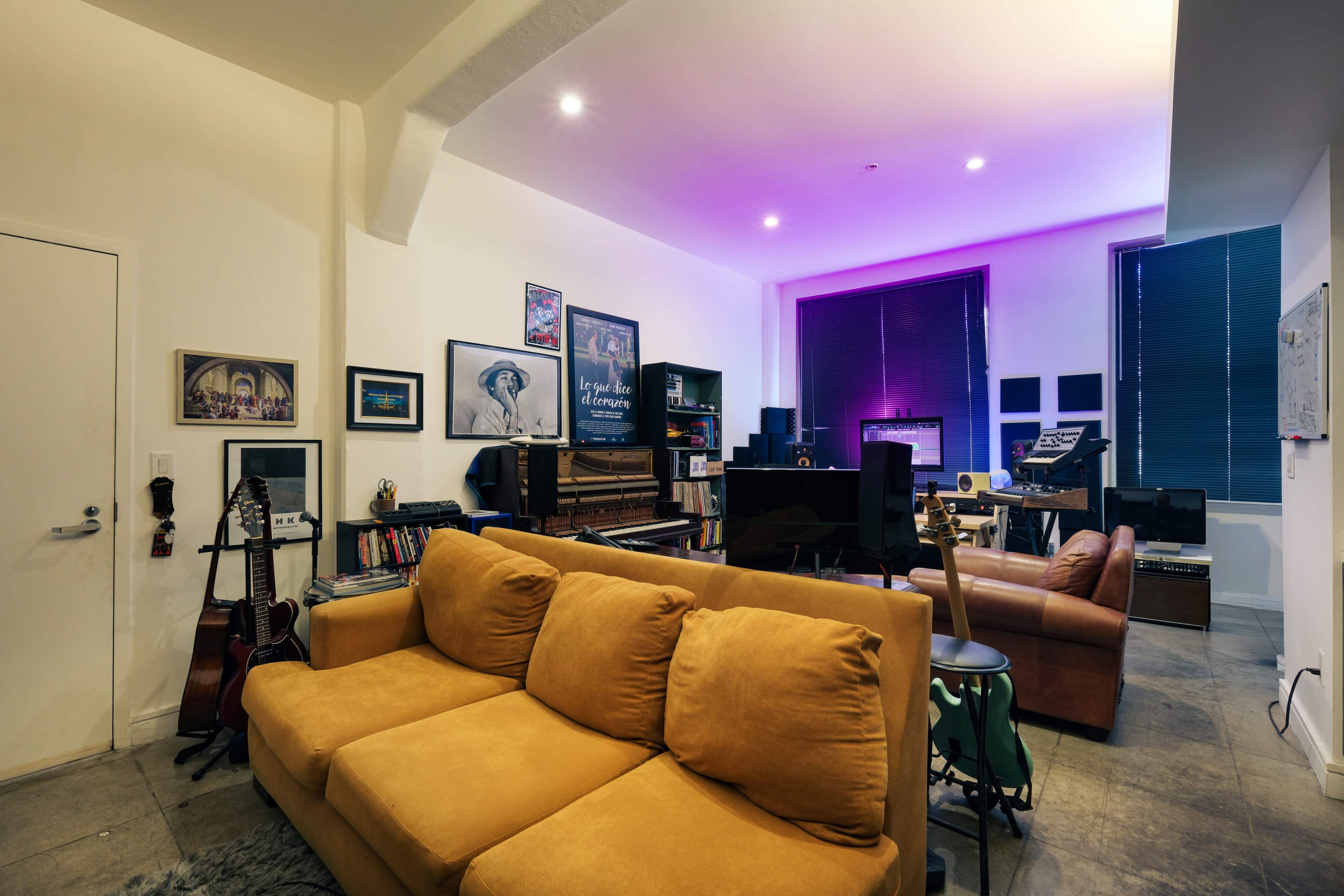 The image shows a cozy living room with a mustard-colored sofa, shelves filled with books and musical instruments, and a workspace featuring a piano and sound equipment under ambient lighting.