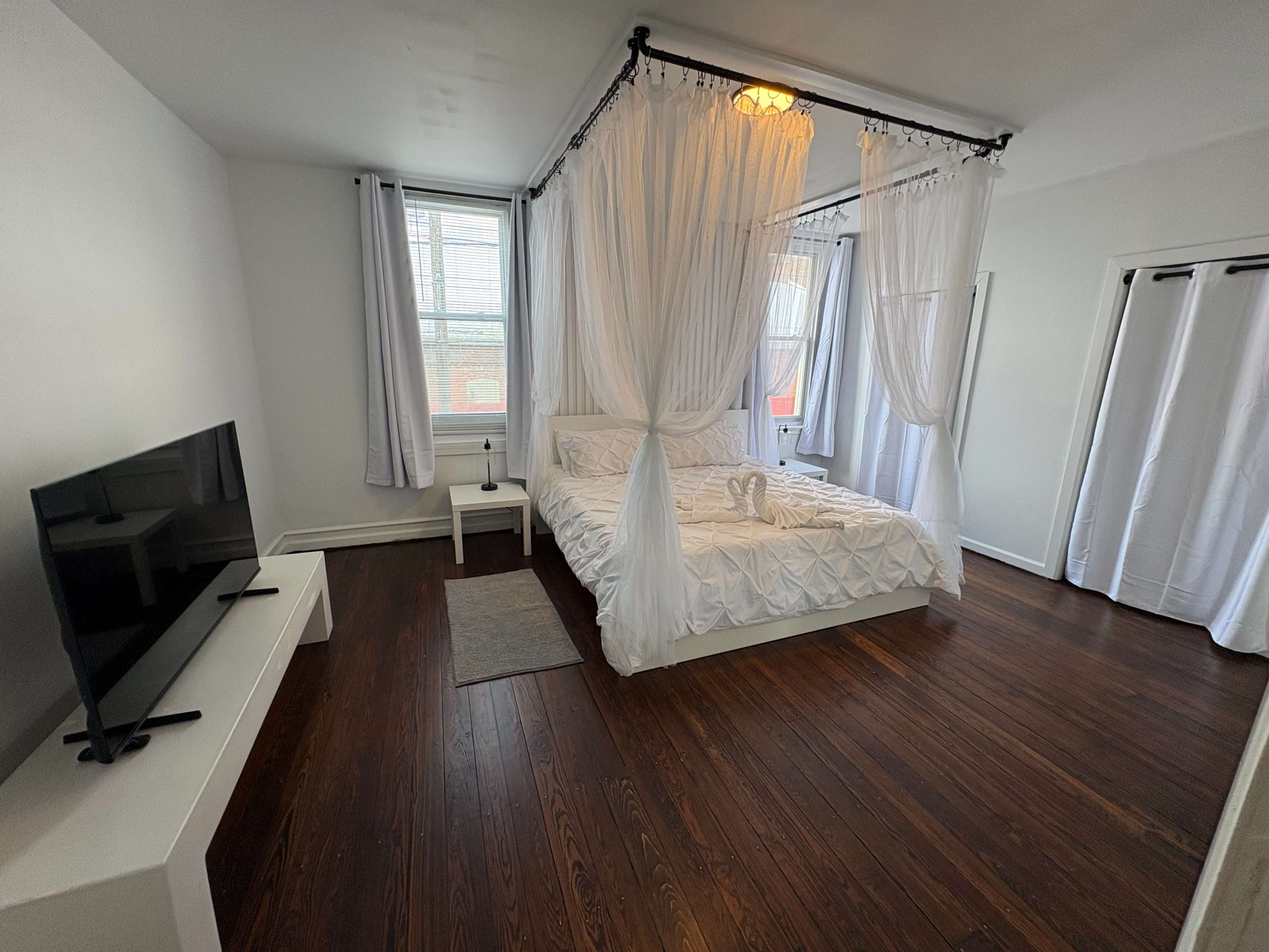 The image shows a bright bedroom with a bed draped in white curtains, a small table, a television on a stand, and wooden floors.