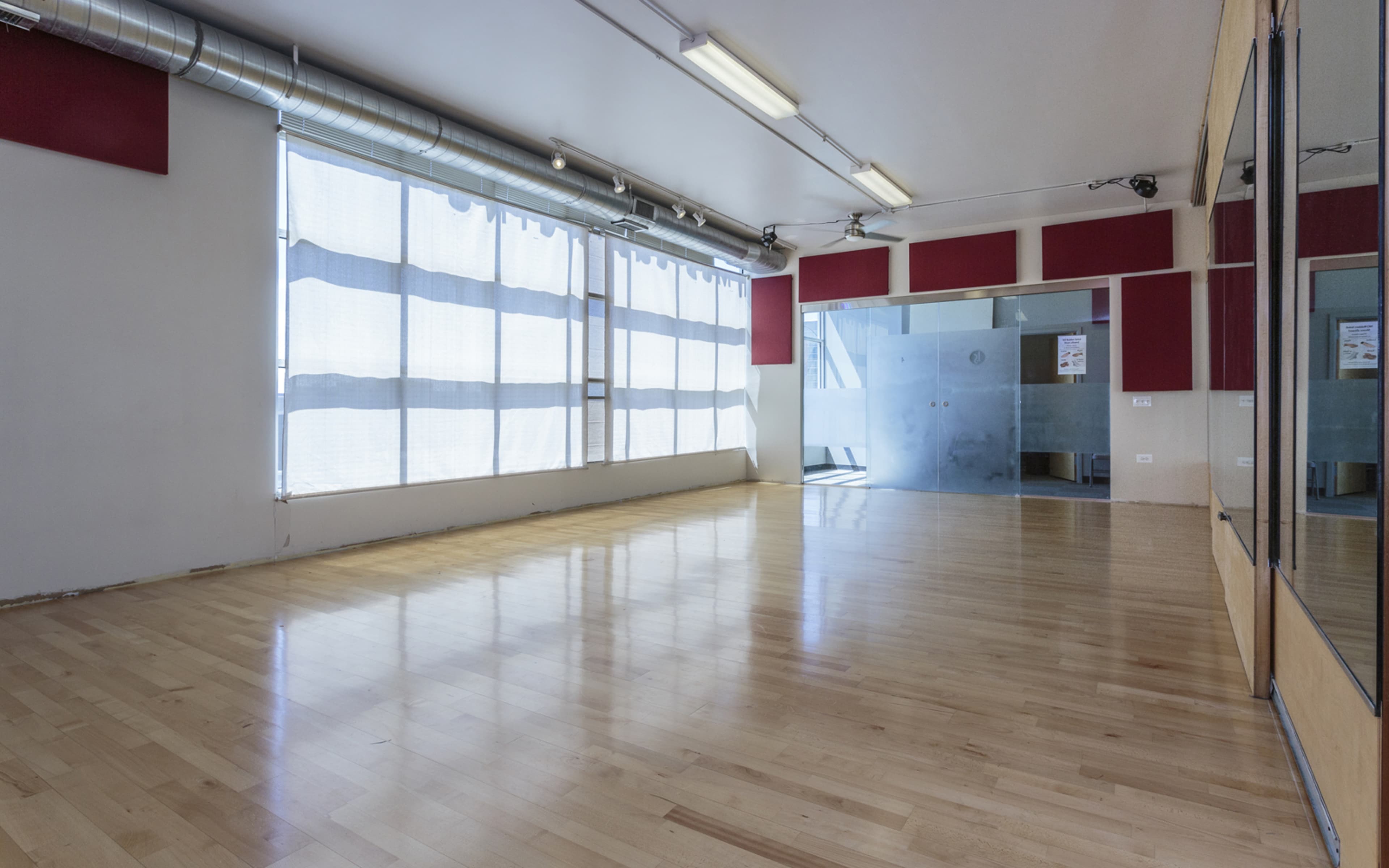An empty, well-lit studio space with wooden floors and large windows covered by sheer curtains.