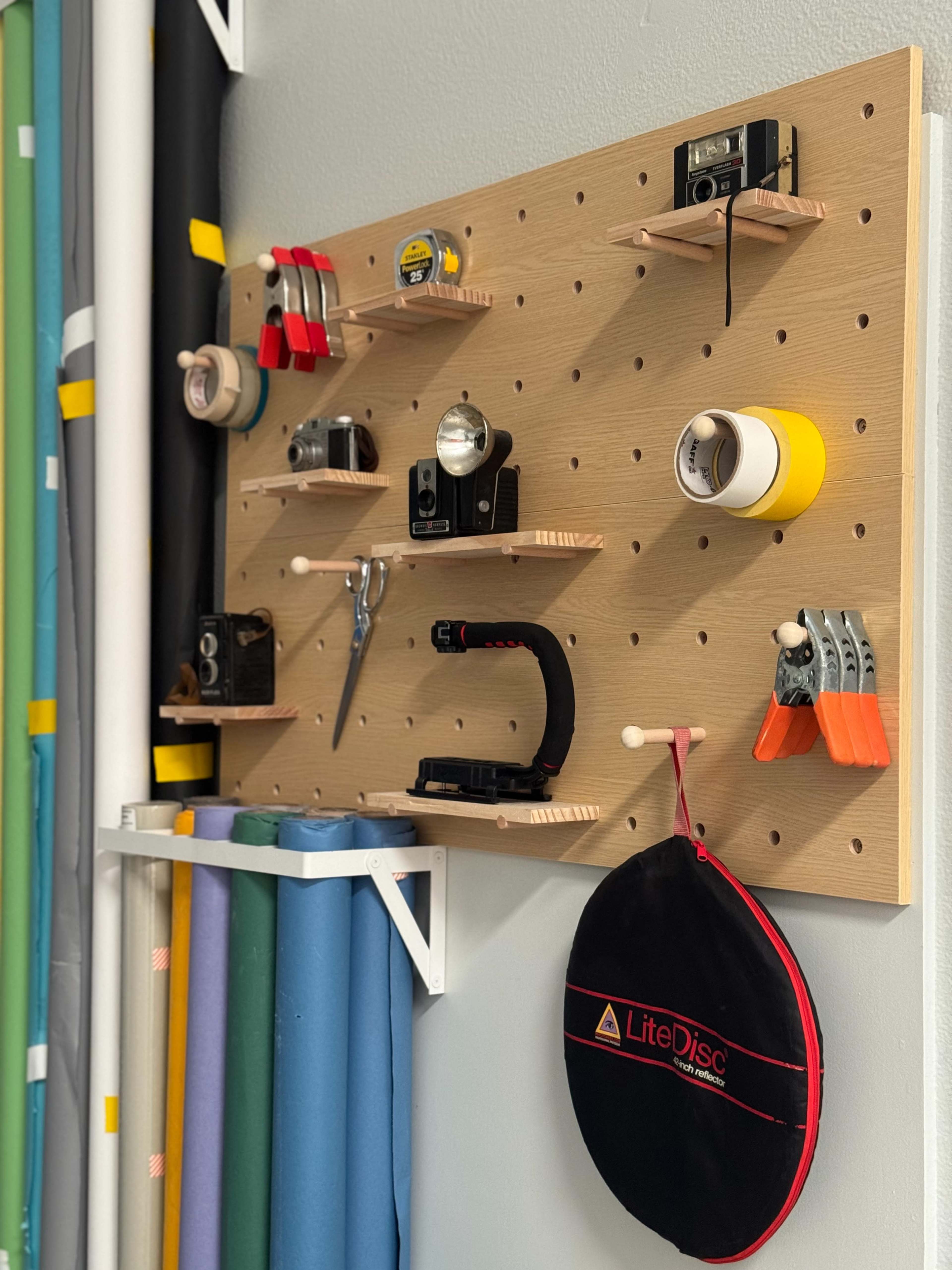 A pegboard is mounted on a wall, displaying various tools, cameras, and a carrying bag, while rolls of colored material are stored below.