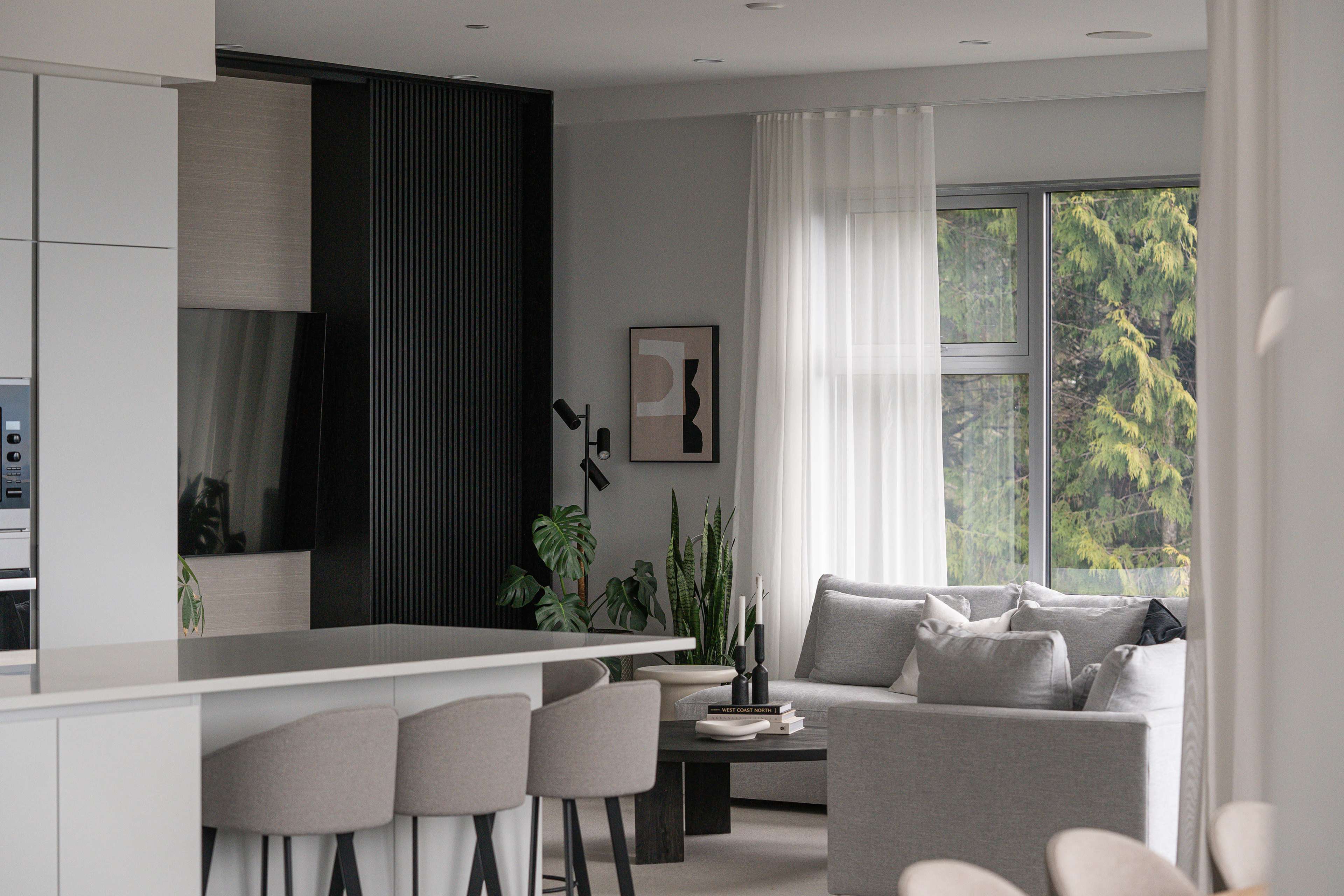 A modern living space features a kitchen area with bar stools, a television mounted on the wall, and a cozy seating area adorned with plants and large windows.