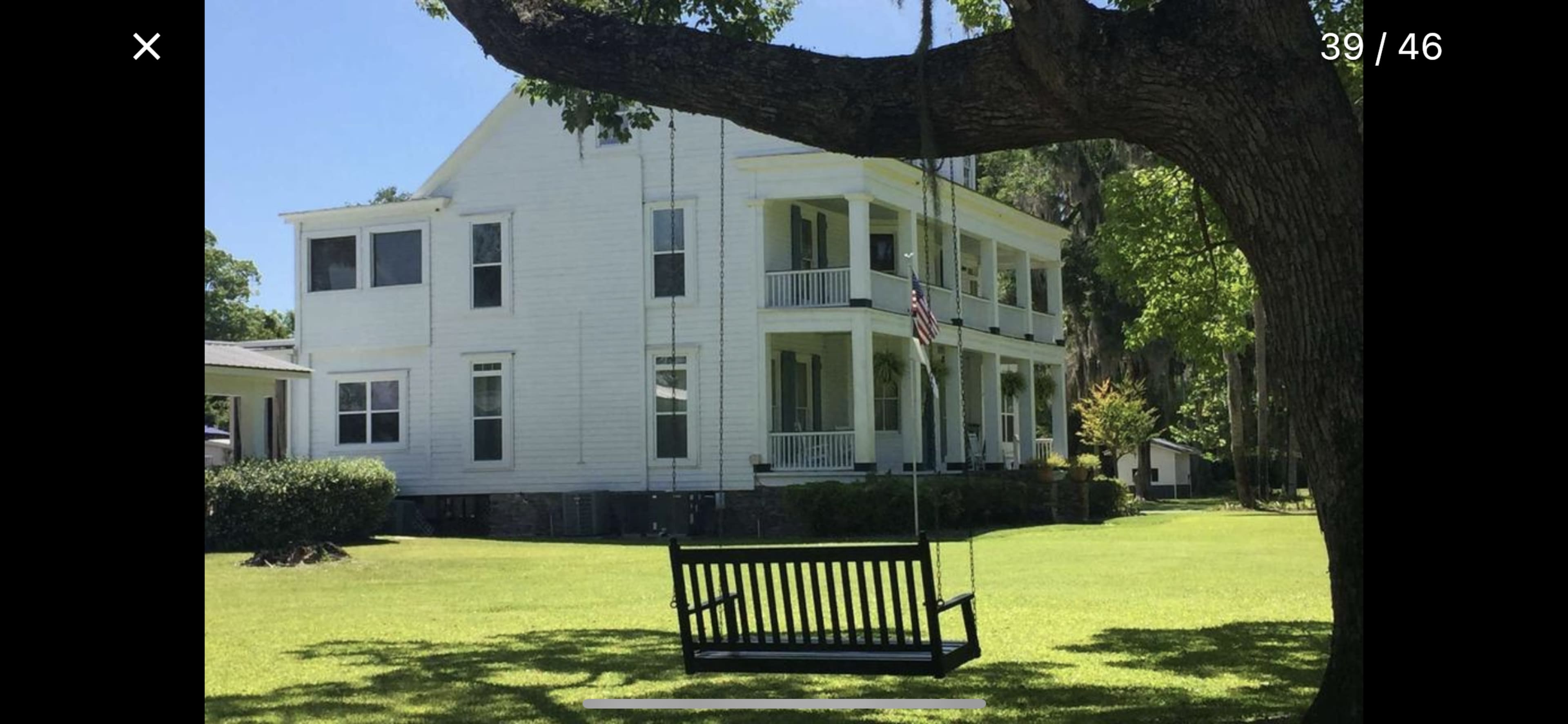 A black wooden swing hangs from a tree in front of a two-story white house surrounded by green grass and trees.