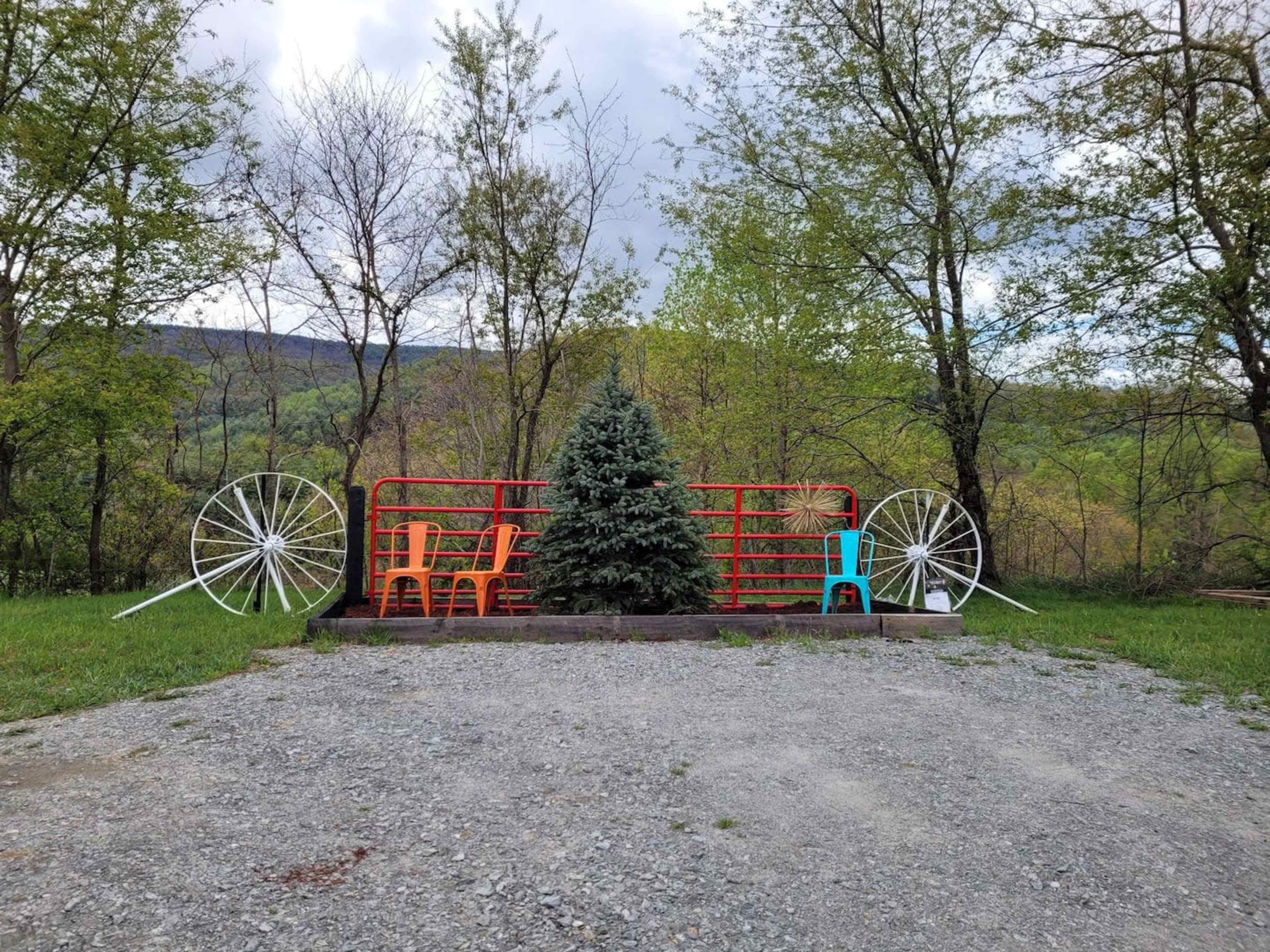 A grassy area with a small red fence, a conifer tree in the center, and brightly colored chairs on either side, framed by large white bicycle wheels.