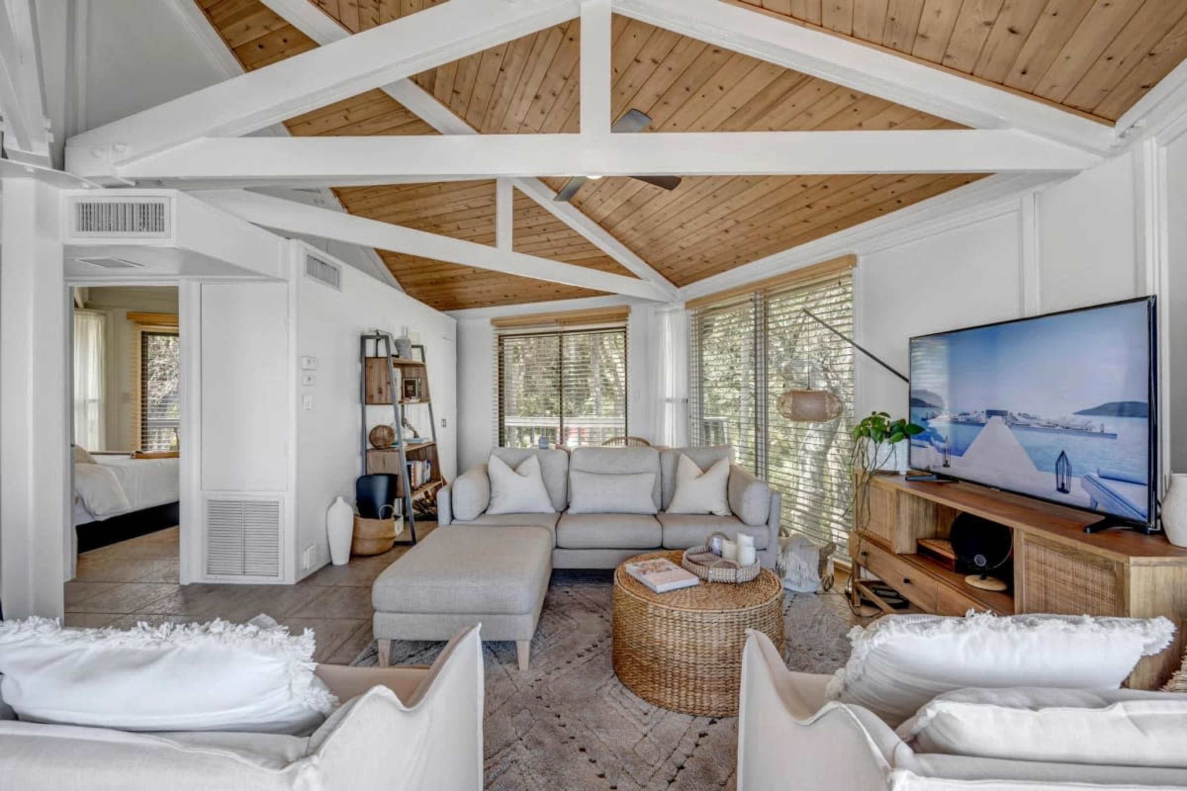 A well-lit living room with a sectional sofa, a coffee table, and a television mounted on the wall, featuring large windows that provide a view of the outside.