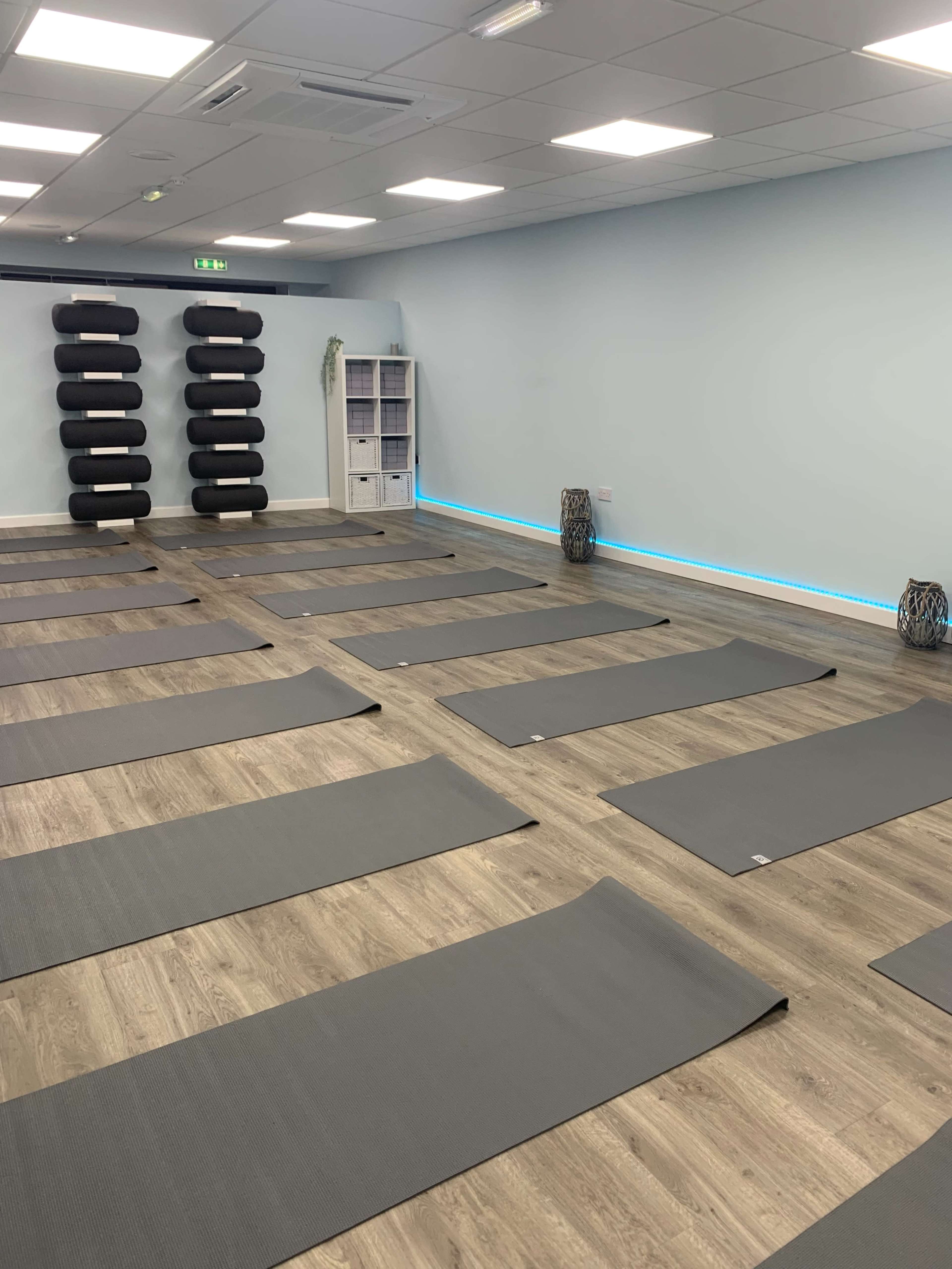 A spacious yoga studio with several gray mats arranged on the wooden floor and black yoga props stacked on a wall.