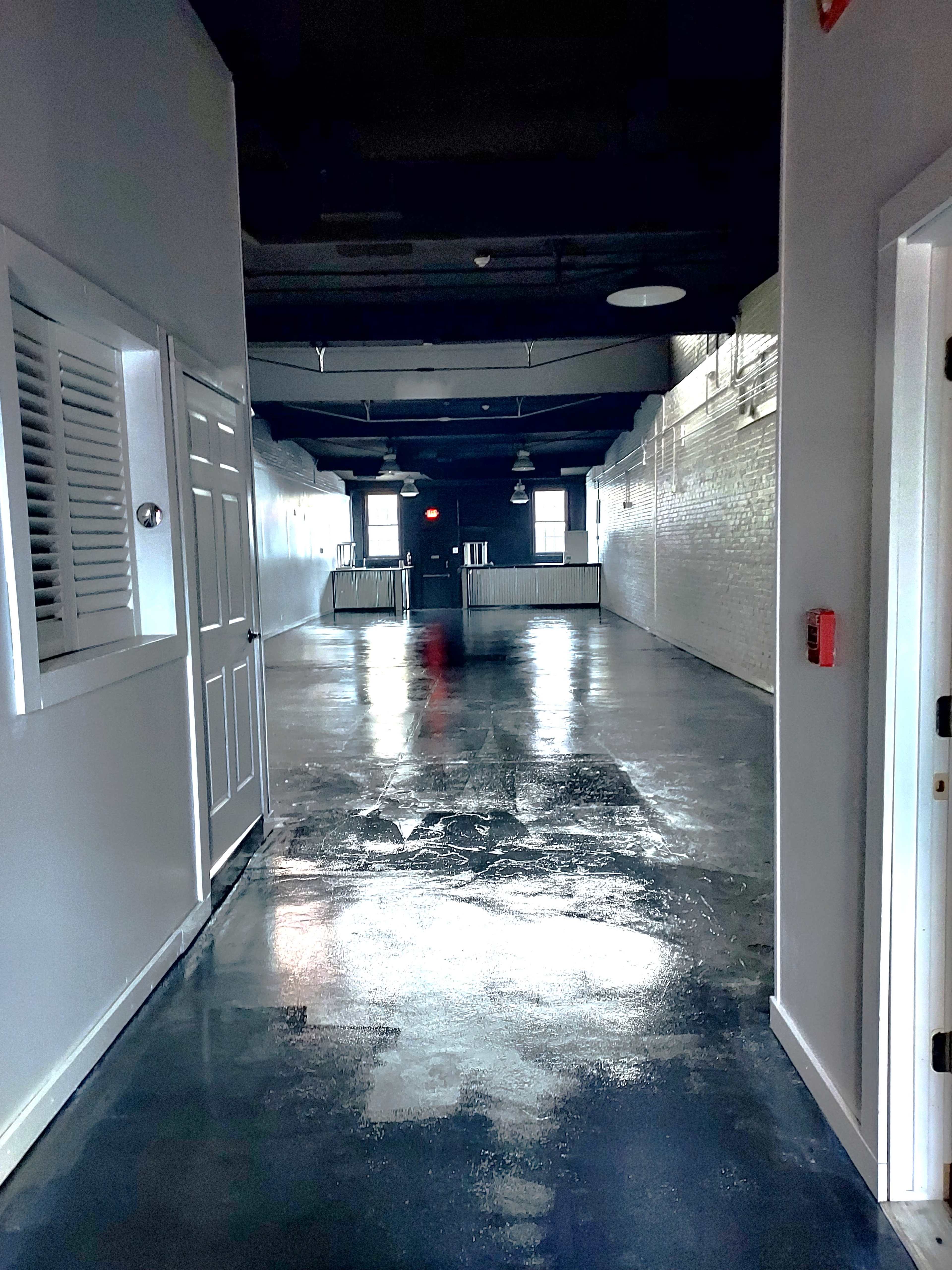 The image shows a long corridor with glossy concrete floors, white brick walls, and doors on either side leading to closed rooms.