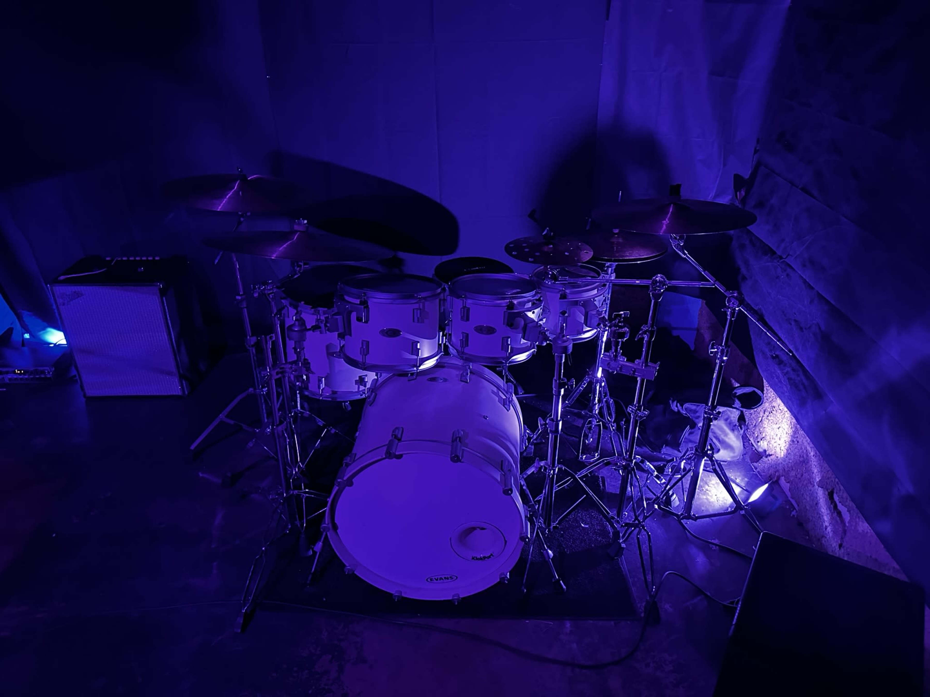 A drum set is positioned in a dimly lit room with blue lighting, accompanied by an amplifier.