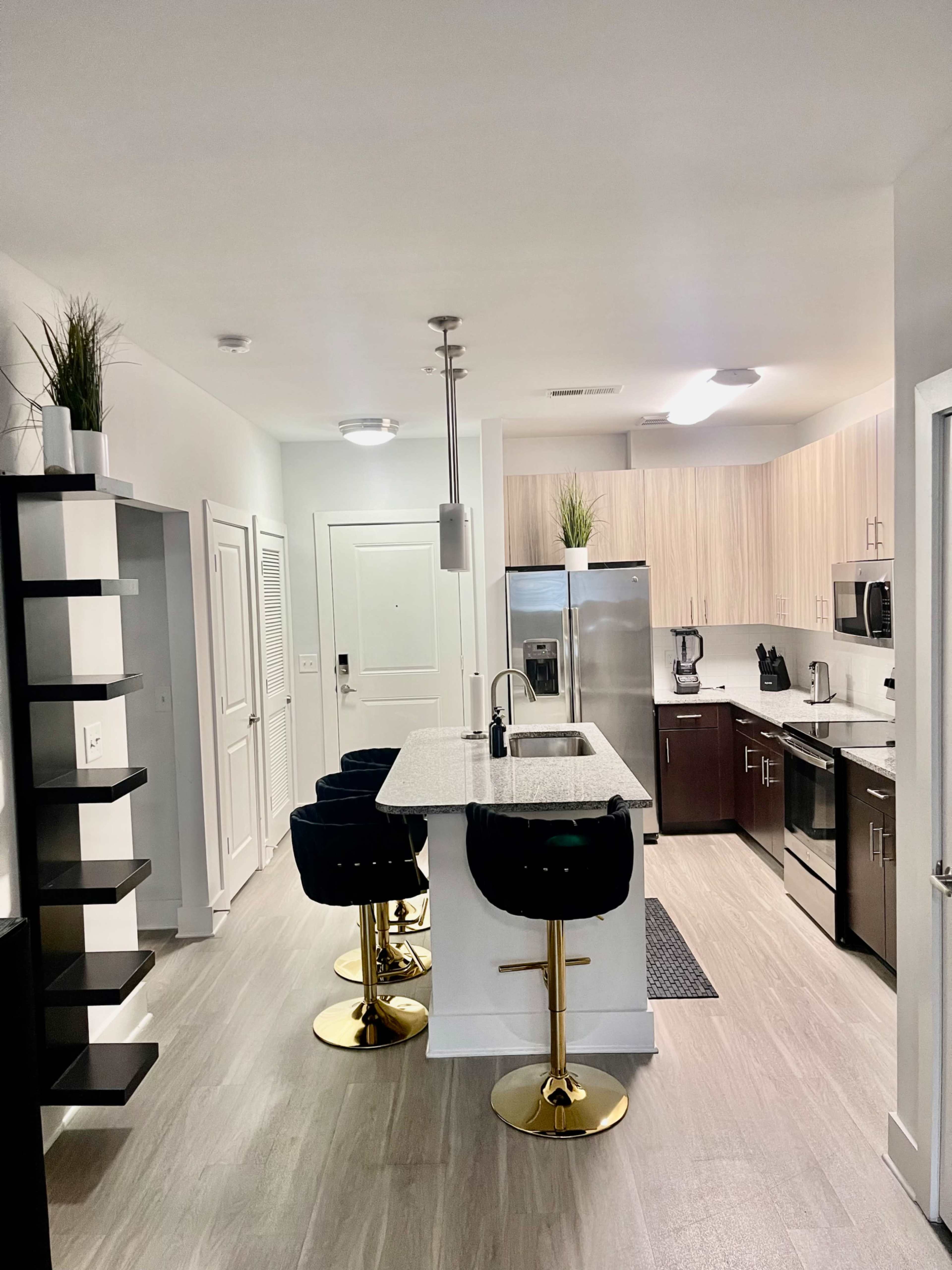 The image shows a modern kitchen featuring a central island with three black bar stools, stainless steel appliances, and light wood cabinetry.