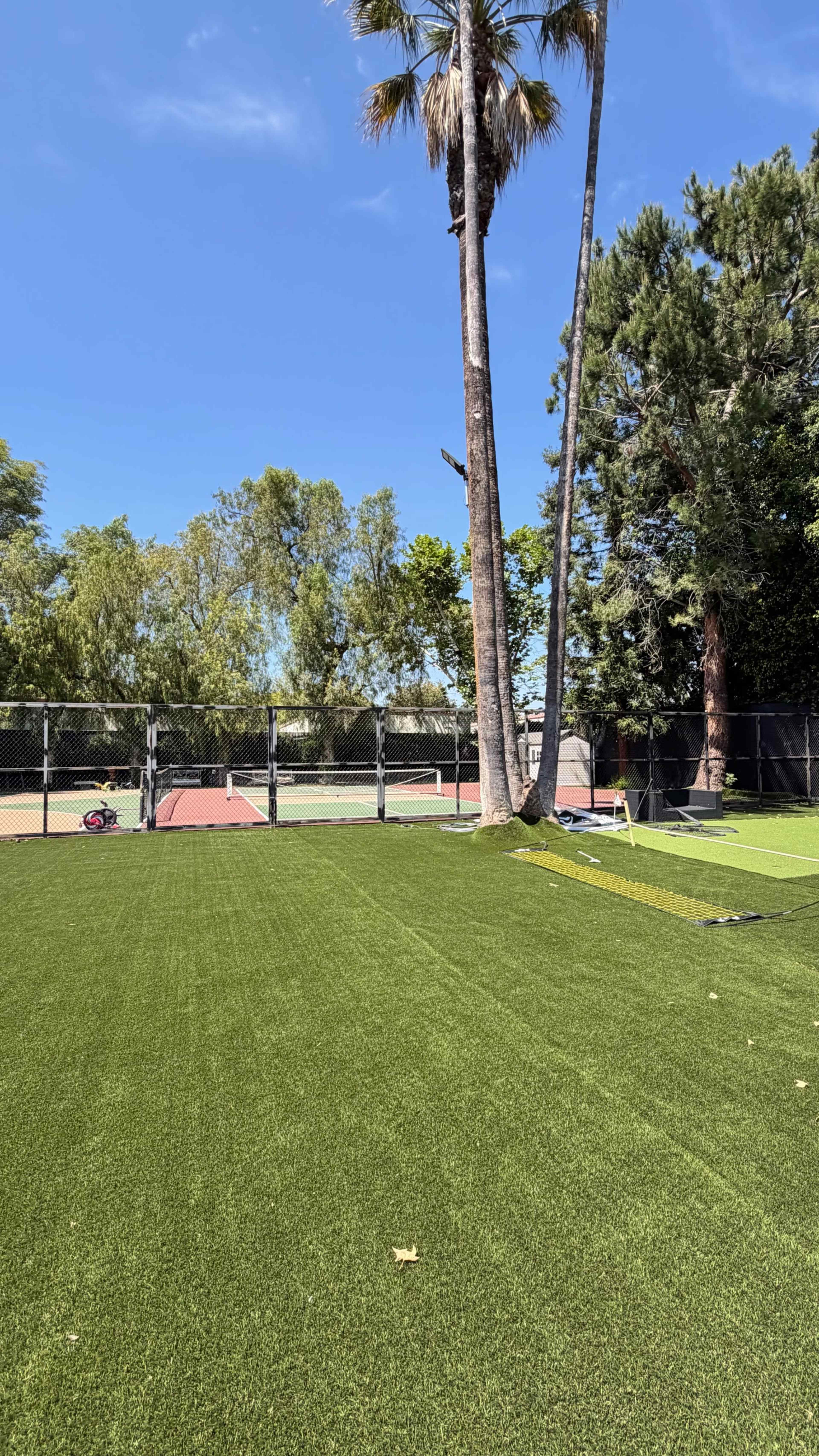 The image shows a well-maintained green artificial turf area adjacent to a tennis court, surrounded by trees and under a clear blue sky.