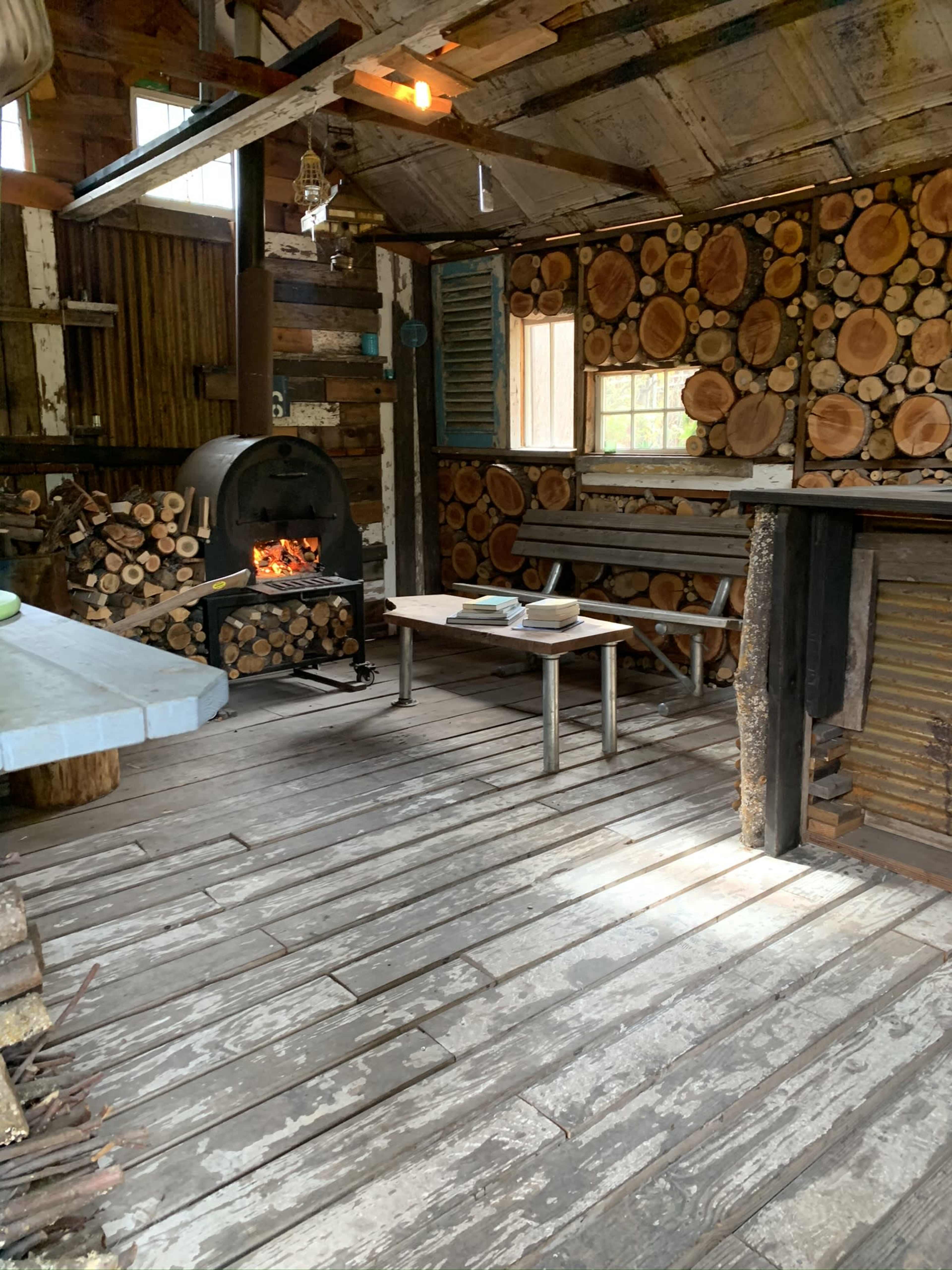 The interior of a rustic cabin features a wooden floor, a stacked log wall, a metal stove with firewood beside it, and a simple table in the center.