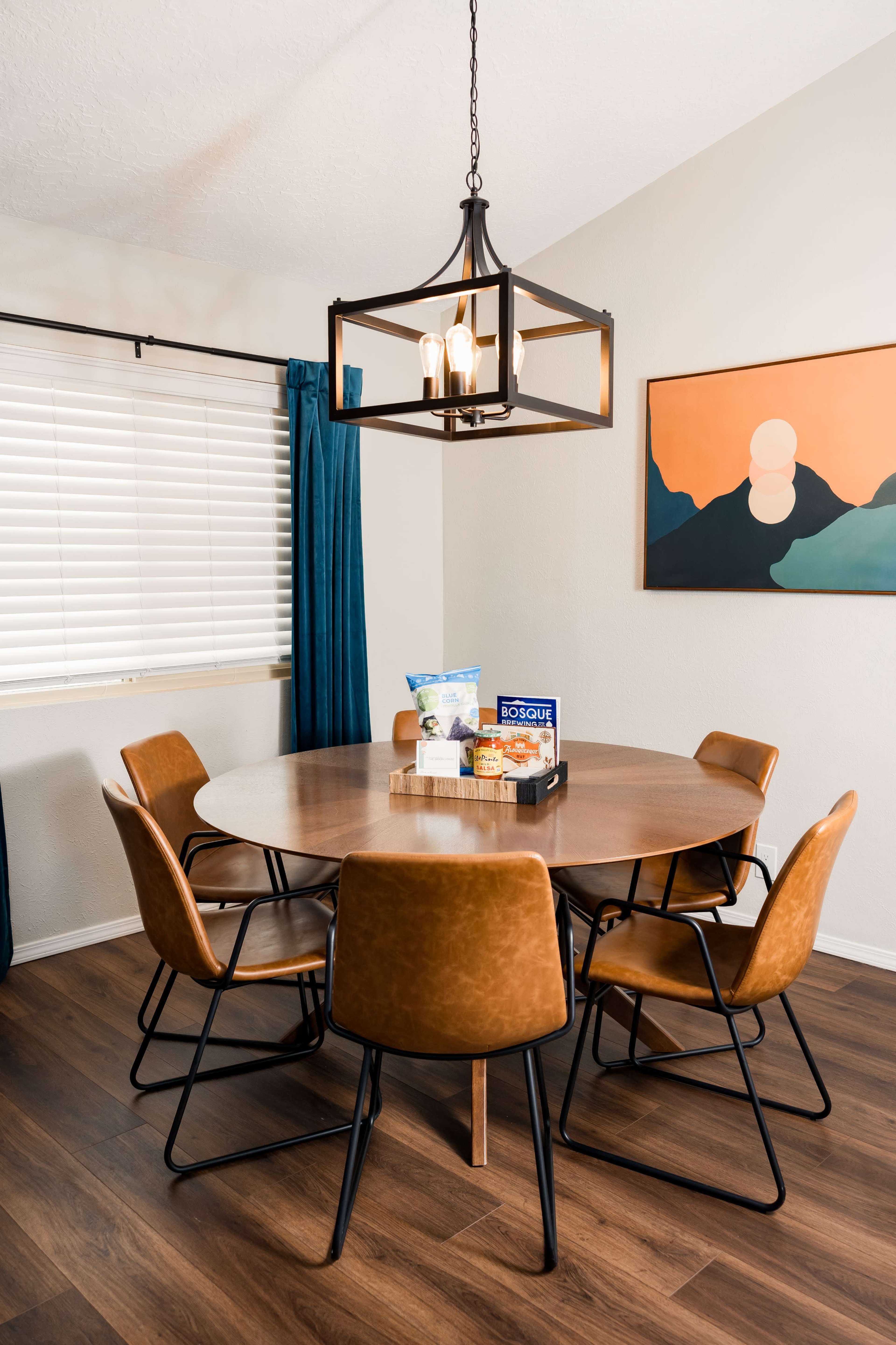 A round wooden dining table surrounded by six leather chairs is illuminated by a pendant light in a well-lit room with a large window and a mountain-themed artwork.