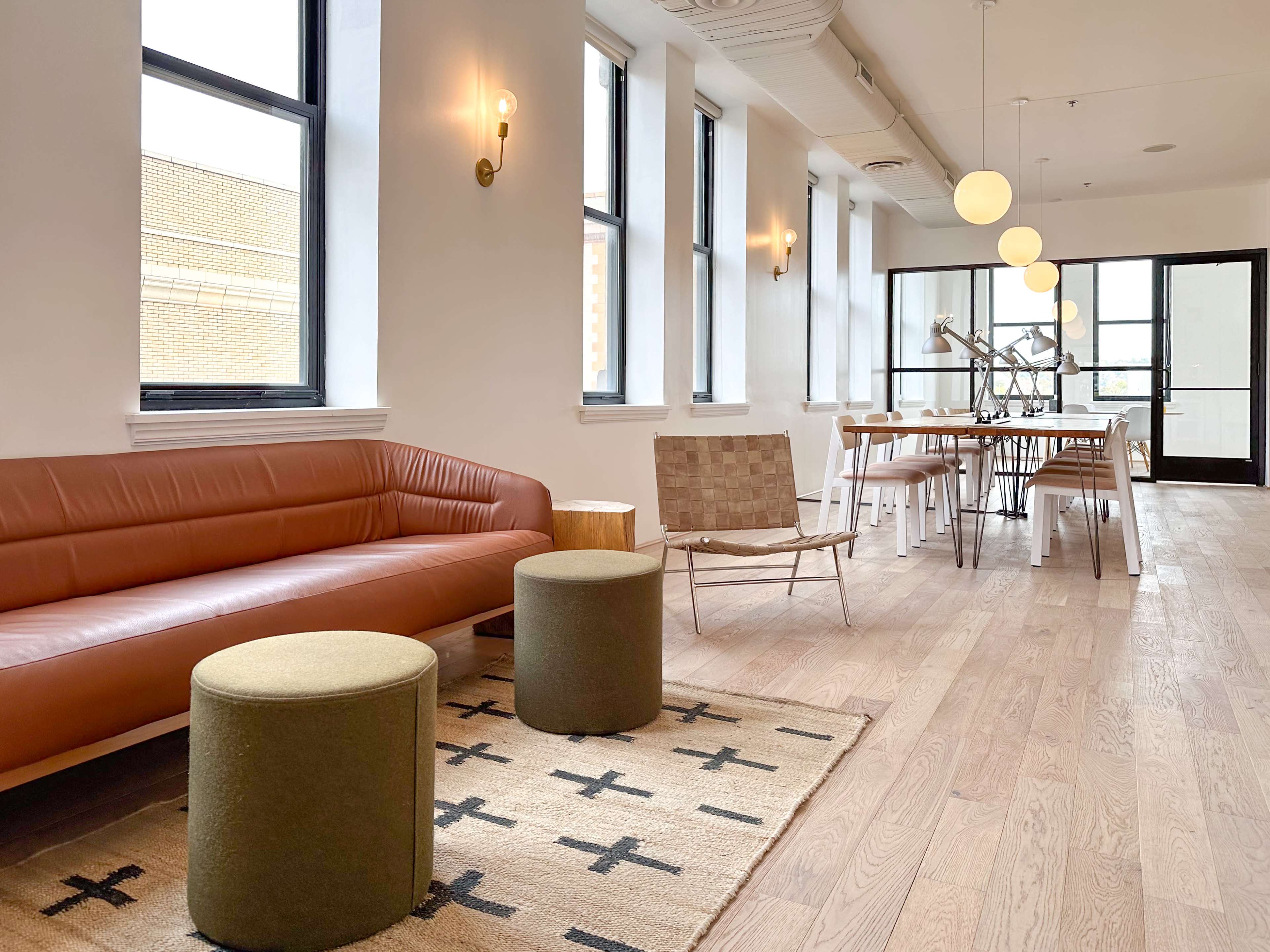 A modern, minimalistic interior featuring a brown leather sofa, two round stools, a woven area rug, a dining table with chairs, and large windows.