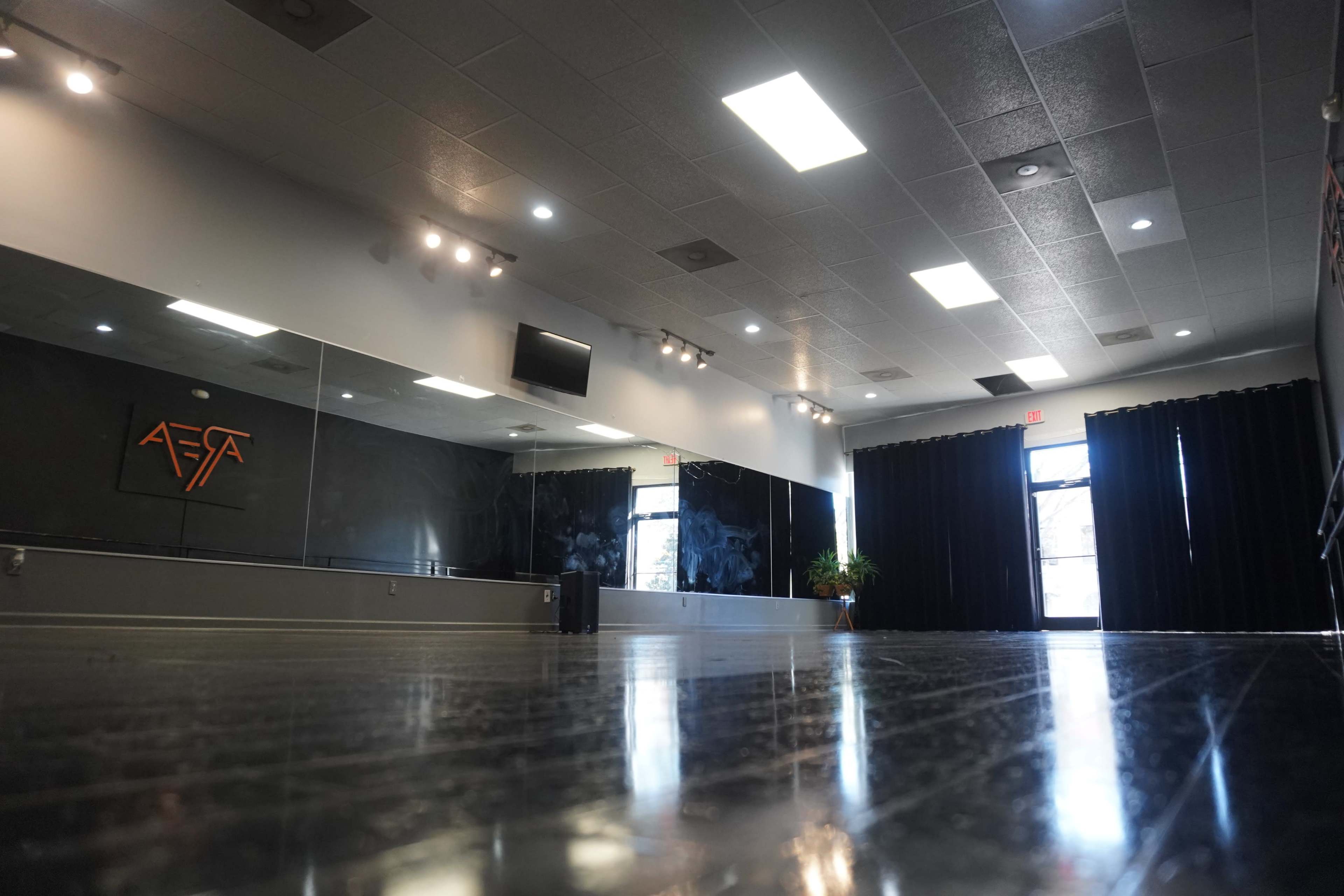 An empty dance studio with a mirrored wall, black curtains, and a wooden floor reflecting overhead lights.
