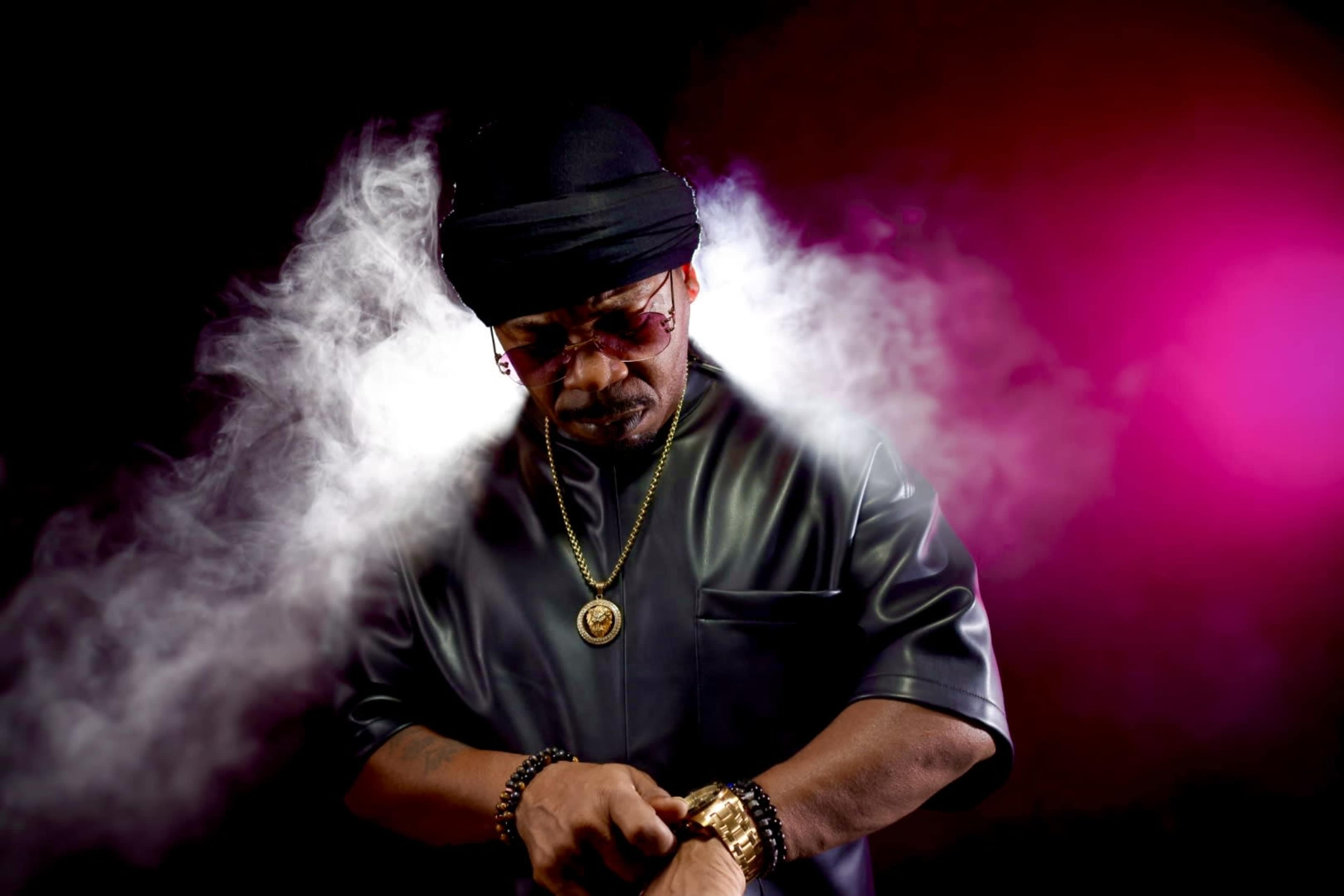 A man in a black outfit and sunglasses adjusts a watch while surrounded by colorful smoke.