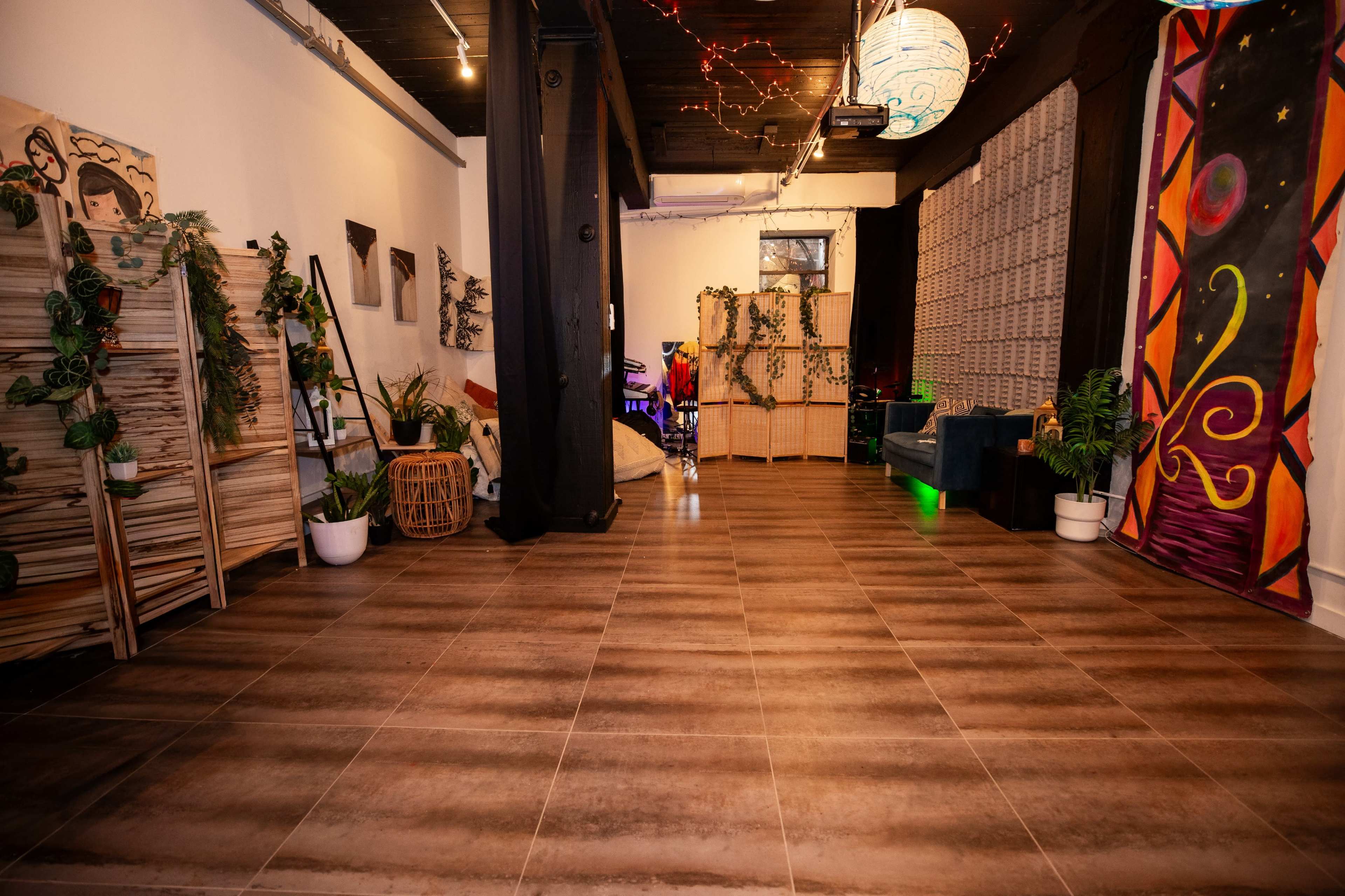 The image shows a stylish interior space featuring wooden flooring, decorative plants, wall art, and soft lighting.