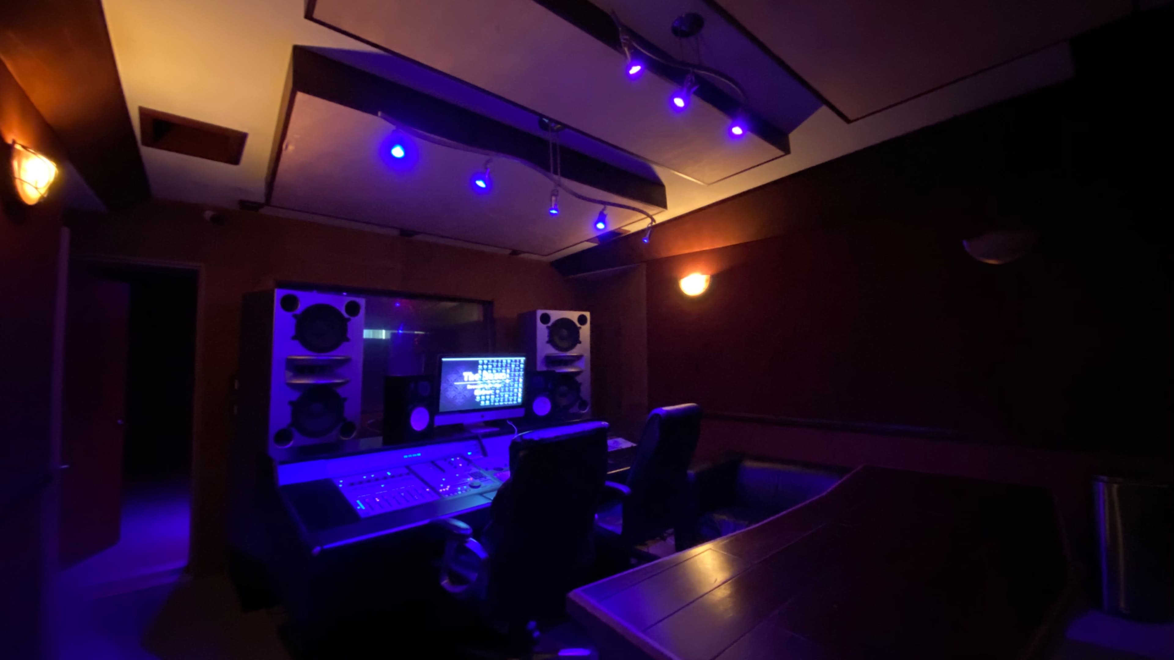 The image shows a dimly lit recording studio with blue lights illuminating a sound mixing console and two chairs facing a large screen.
