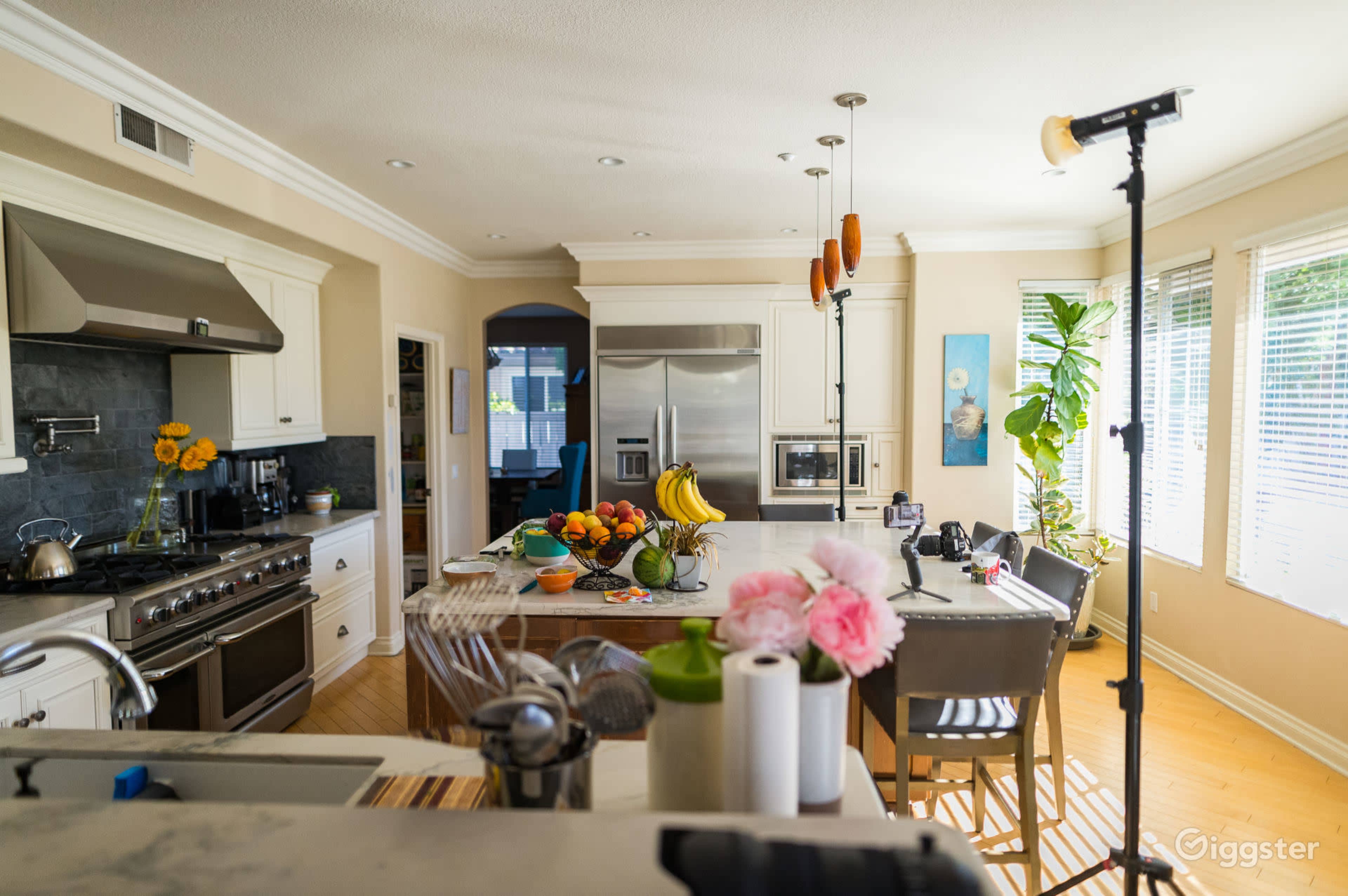 A well-lit kitchen features stainless steel appliances, a large island with fresh fruit, and a counter adorned with flowers and kitchen utensils.